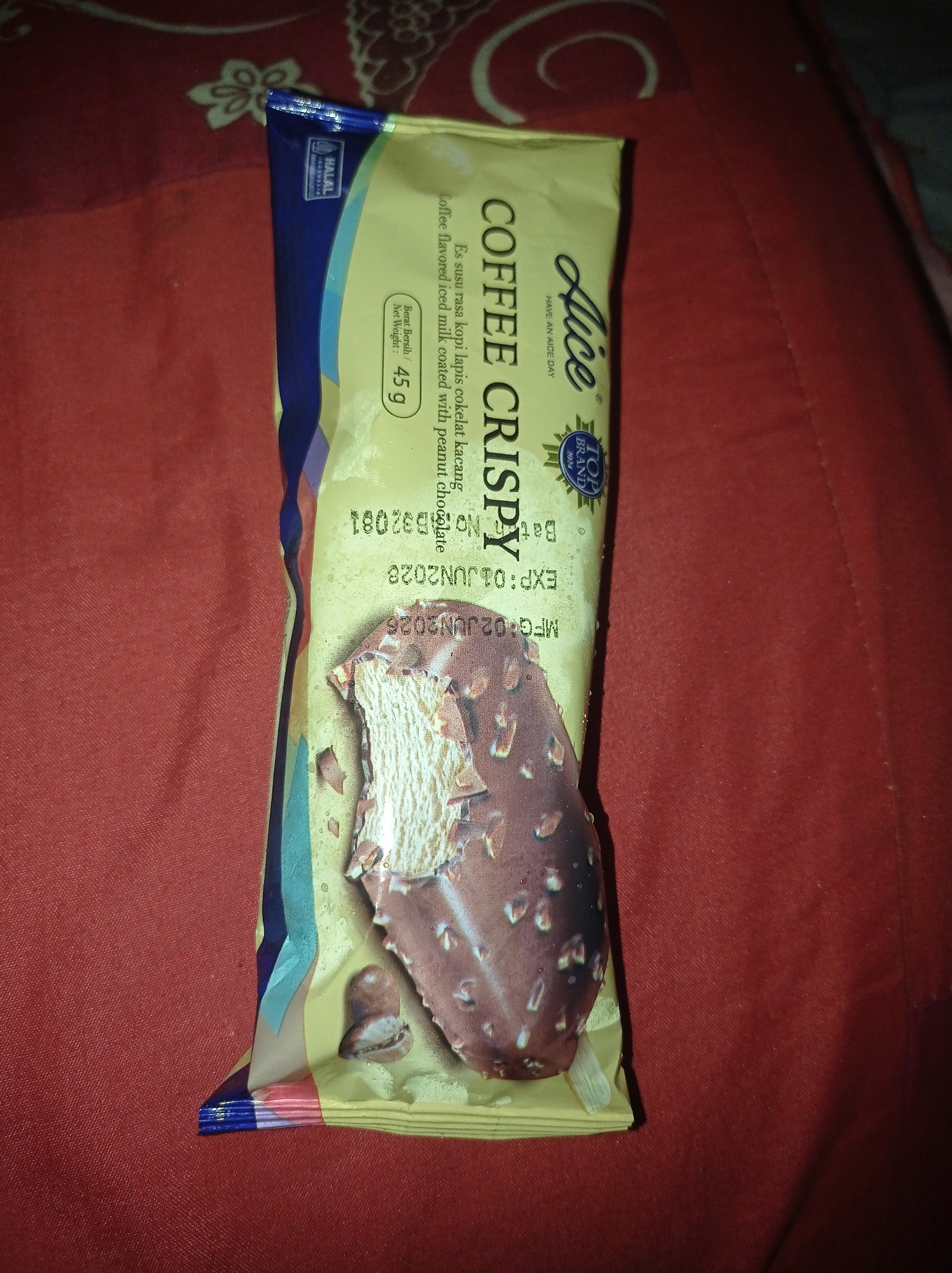 The image shows a wrapper for an ice cream bar on a red textured surface. The wrapper is primarily yellow and features text and graphics related to the product. The ice cream bar itself is partially visible, appearing to be chocolate-coated with nuts and filled with a creamy interior. The wrapper has text indicating the brand "Alice", a flavor described as "COFFEE CRISPY", and production details like "MFG:02JUN2026" and "EXP:01JUN2026". There is also a "HALAL" logo in the upper left corner. The red surface beneath the wrapper appears to be fabric, possibly a bedsheet or blanket, with some subtle patterned texture. The image is taken from a slightly elevated angle, with the wrapper positioned vertically. There are no people or specific environmental details of a city like Sumbernadi, Indonesia visible in the image.