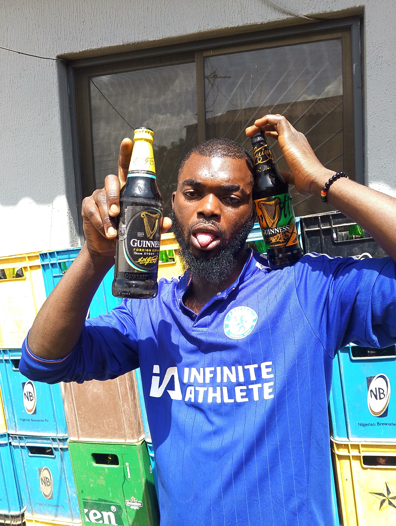 A man with a beard and a blue shirt is holding up two bottles of Guinness beer, one in each hand, over his head. He is sticking his tongue out at the camera, and his eyes are closed. The man is standing in front of a stack of plastic crates, some of which are blue and yellow, and others are green. Some of the crates have the "NB" logo, which stands for Nigerian Breweries Plc, and the text "Ken" is visible on one of the green crates. The background shows a building with a window. The scene appears to be outdoors, likely during the daytime, given the bright sunlight and shadows. The man's expression and pose suggest a playful and casual mood.