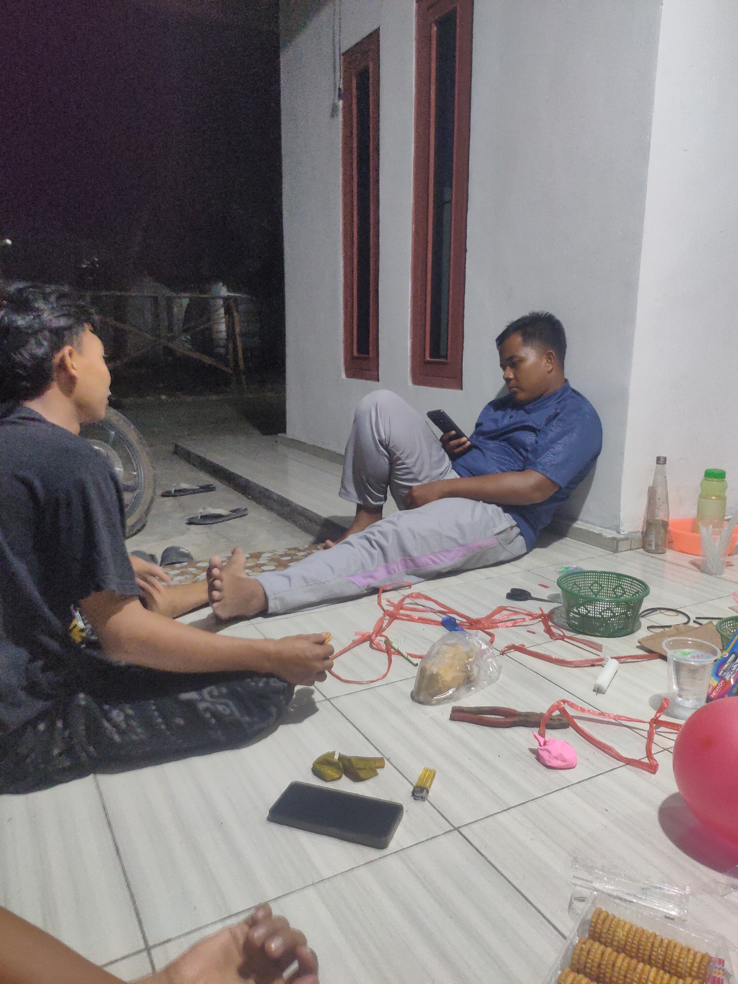 Two men are seated on the tiled floor outside a building with white walls and dark-framed windows. The man on the left, wearing a black t-shirt, faces the man on the right and is seen from the back. The man on the right, wearing a blue short-sleeved shirt and grey pants, is leaning against the wall and looking at his smartphone. The ground is cluttered with various items, including a green basket, red plastic strapping, a plastic bag containing food, a lighter, a pink object, scissors, and a large pink balloon. A pair of flip-flops are visible on the floor near the man on the left. The lighting suggests it is nighttime. The scene appears to be the exterior of a residential building in Pematang Kulim, Indonesia.