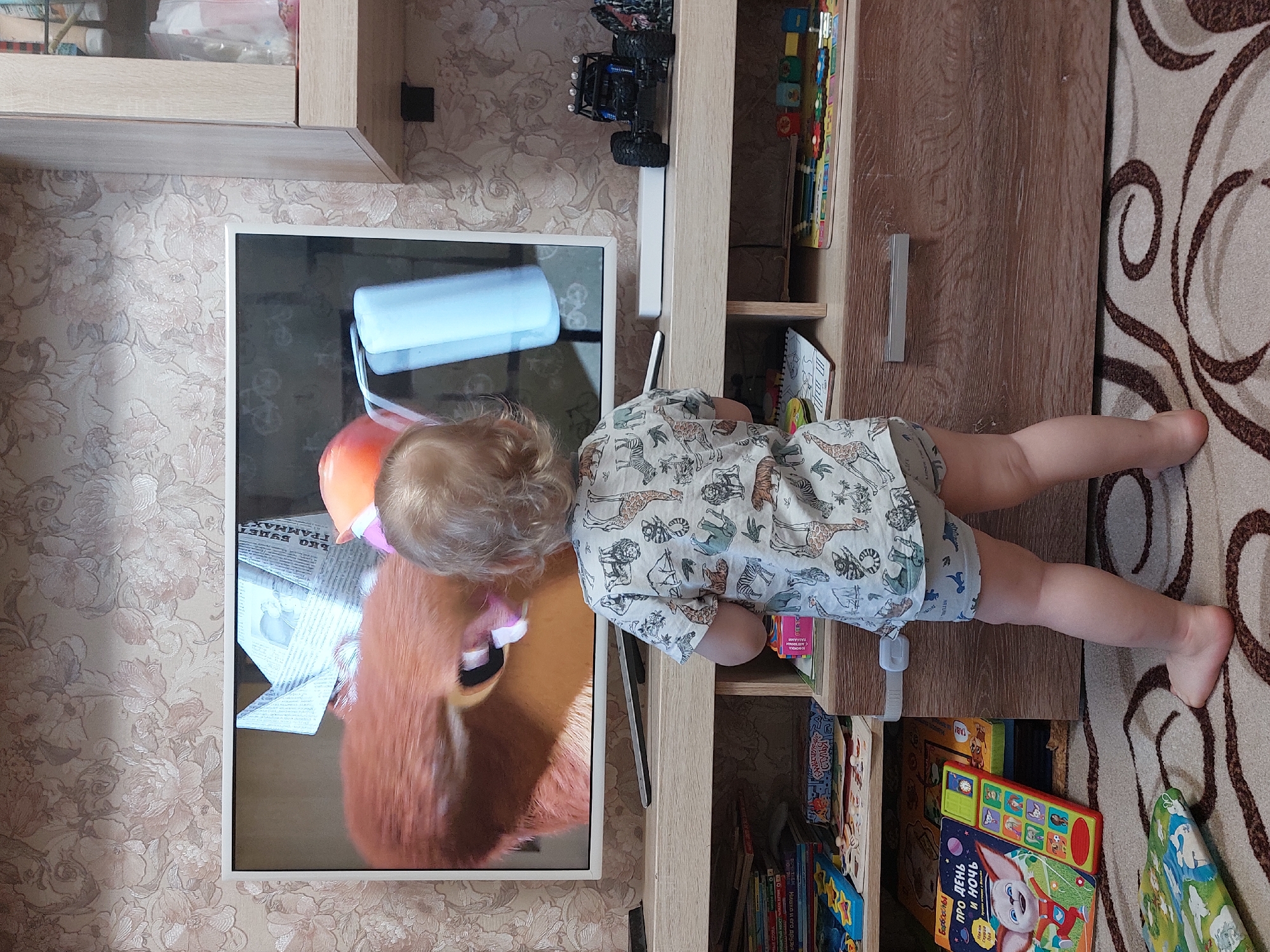 The image shows a child leaning over the edge of a wooden cabinet with books on its shelves. The child is wearing a shirt with a giraffe and lion print. The child's legs are on a rug. A TV showing a cartoon of a large orange plush animal is on the cabinet. The TV rests against a wallpaper with a floral pattern. The Russian text on the book "ПРО ДЕНЬ И НОЧЬ", on the newspaper "Чайковский", and the filename suggests the location may be in Chaikovsky, Russia.