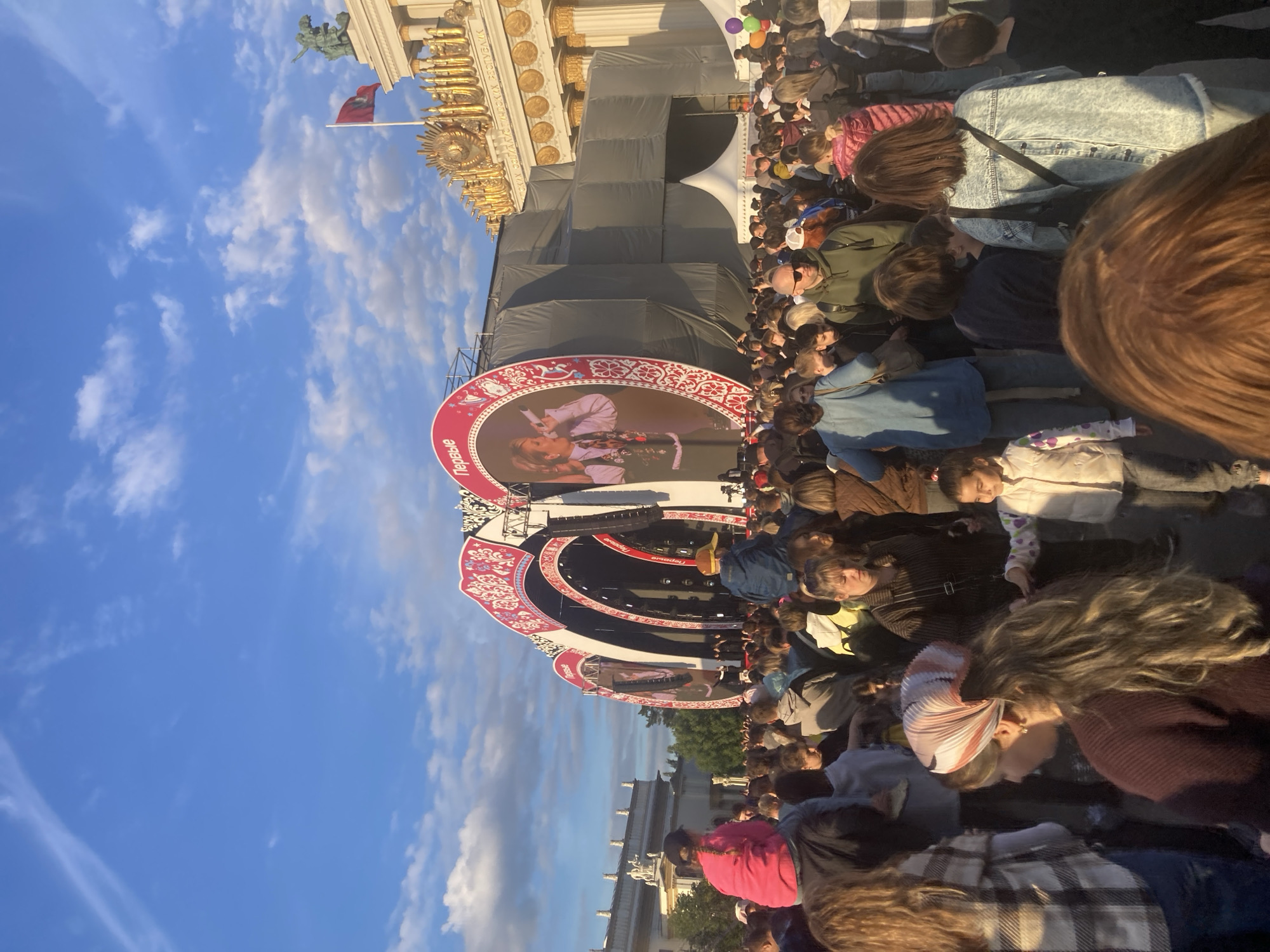The image depicts a large public gathering, likely a concert or festival, taking place outdoors in what appears to be late afternoon or early evening under a clear blue sky with scattered white clouds. A substantial crowd of people, comprising adults and some children, faces a prominent stage.
The stage features multiple large, circular screens framed with ornate red and white patterns, reminiscent of traditional Russian folk art. On the screens, a female performer with blonde hair, wearing a white blouse and a patterned skirt, is shown singing into a microphone. The Russian word "Первые" (Pervye, meaning "The First") is visible multiple times on the decorative frames of these screens.
In the background, a grand, ornate building with golden architectural elements and columns is visible. A red flag featuring a golden emblem, possibly a double-headed eagle, flies from a flagpole attached to this building. Faintly visible Cyrillic text on the building's frieze includes parts of "ВСЕСОЮЗНАЯ ВЫСТАВКА ХОЗЯЙСТВА" (All-Union Exhibition of Economic Achievements), indicating the location is VDNKh (Exhibition of Achievements of National Economy) in Moscow, Russia.
The crowd appears to be quietly observing the performance, with most individuals seen from behind or from the side. The overall scene suggests a cultural event attracting a considerable audience, set against an iconic architectural backdrop under pleasant weather conditions.