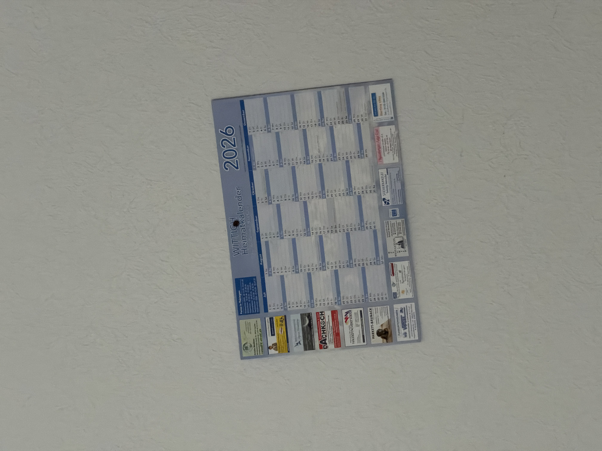 The image shows a wall calendar for the year 2026, tilted slightly and viewed from an upward angle. The calendar itself is a rectangular poster with a blue and white color scheme. It features a grid layout for each month, with days of the week and corresponding dates clearly visible. The months displayed range from July to December.
Prominently featured on the left side of the calendar is the text "WITTIGH Heimatkalender 2026", indicating it is a local calendar for a place called Wittigh, and it is for the year 2026.
Advertisements from various businesses are attached to the bottom and right sides of the calendar. These include:
- "STEUERKANZLEI" (Tax Consultancy) with an address and contact information.
- "DACHKOCH" (Roofing) with a prominent logo and phone number.
- "PARKETT-BAUNACK" (Parquet Flooring) with a smaller advertisement.
- A business related to "PUHKUNTERNEHMEN" (Puhk Enterprises).
- An advertisement for "Mertina Ulies" with a phone number.
- A small yellow advertisement with "4th" and some German text.
- Another advertisement with "Fenster in Thüringen" (Windows in Thuringia).
The wall appears to be textured, possibly painted with a rough plaster finish, in a neutral light color. The lighting suggests it might be indoors, with the light source casting subtle shadows. There are no people visible in the image, and the focus is entirely on the calendar and its accompanying advertisements. The overall impression is of a functional item displayed in a practical setting, likely an office or a home.