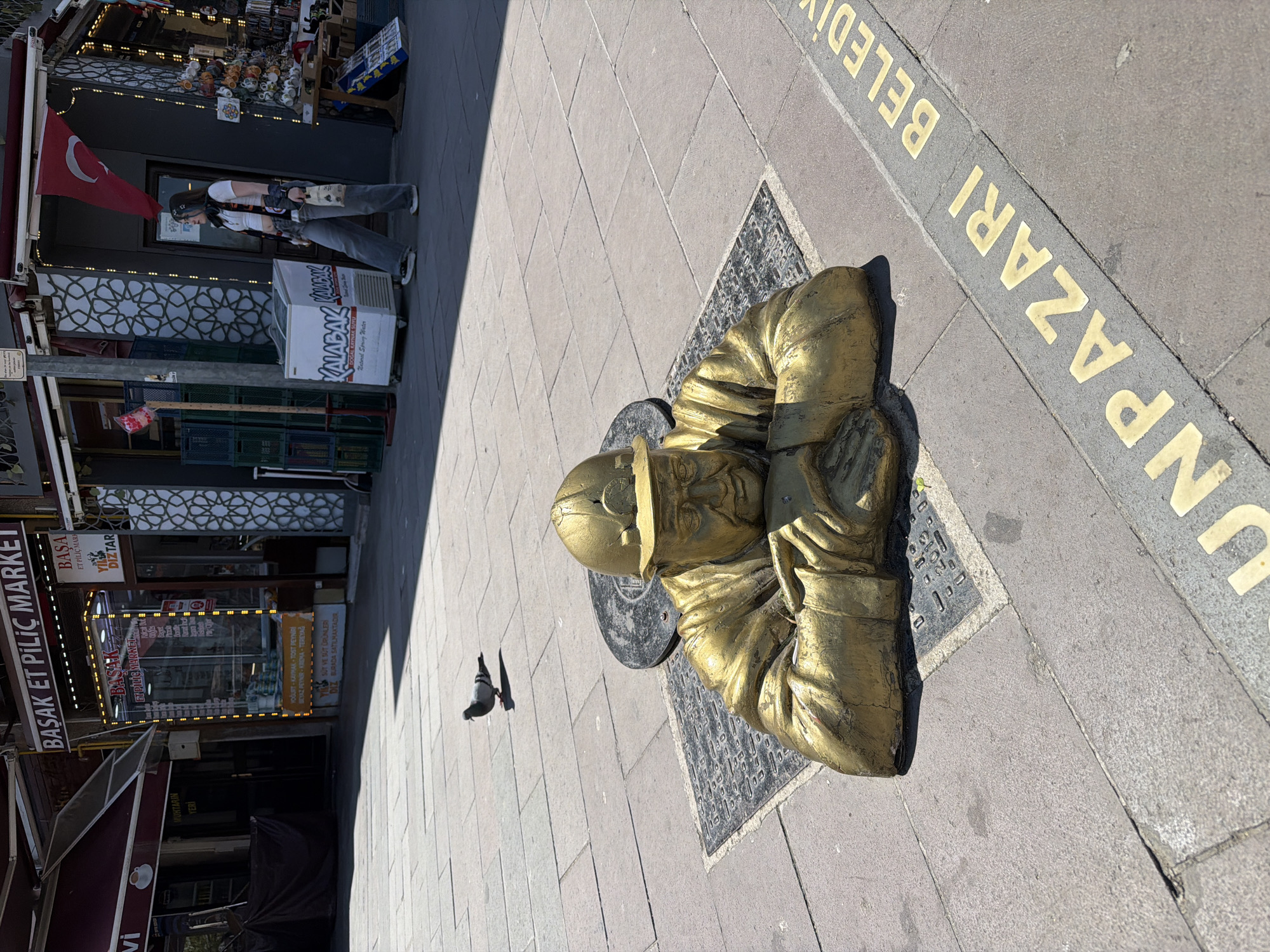 The image displays a vibrant street scene in Eskisehir, Turkey, illuminated by bright daylight, casting distinct shadows. The central subject is a golden statue of an elderly man, possibly depicting a traditional craftsman or vendor, positioned low to the ground on a black-patterned base. He appears to be kneeling or sitting, with his hands resting on his knees.
The statue is situated on a paved sidewalk, adjacent to a strip of golden lettering embedded in the ground that reads "UNPAZARI BELEDİ," likely part of "Odunpazarı Belediyesi," the local municipality. To the left of the statue, a pigeon pecks at the ground.
Along the street, several storefronts line the buildings. A red Turkish flag, bearing a white crescent and star, is prominently displayed above one shop entrance. Near this shop, a person stands by a white cooler, seemingly engrossed in their phone or another handheld device. The cooler is labeled "KALABAK DOĞAL KAYNAK SUYU" (Kalabak Natural Spring Water) and "External Spring Water." The building features a decorative white lattice pattern on its facade.
Further along, another shop, "BAŞAK ET PİLİÇ MARKET" (Başak Meat and Poultry Market), is visible with brightly lit signage. Other signs detail available products and services: "MUHTARIN YERI" (Headman's Place/Office) and a list of dairy items including "YOĞURT-KAYMAK TOST PEYNİRİ BEYAZ PEYNİR-KREMA TEREYAĞ" (yogurt, clotted cream, toast cheese, white cheese, cream, butter), explicitly stating "SÜT VE SÜT ÜRÜNLERİ BURADA SATILMAKTADIR" (milk and dairy products are sold here). The scene captures a slice of daily life in a lively urban setting.