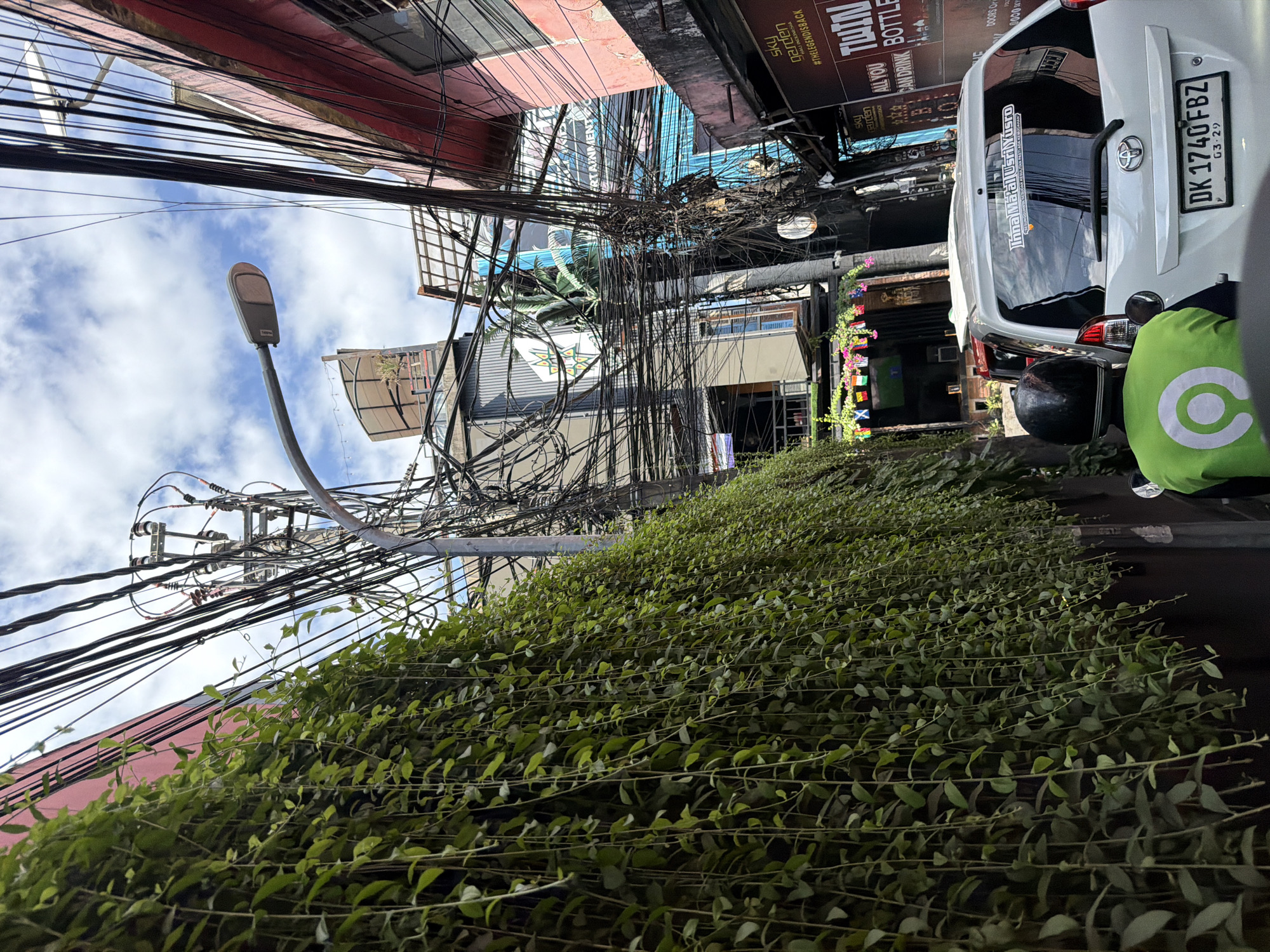 The image depicts a narrow street in Kuta, Indonesia, densely packed with overhead electrical wires and lush green hanging vines cascading down a red-painted building on the left. The sky is partly cloudy. In the foreground, a white car with a license plate reading "DK 1740FBZ" is visible, and a person wearing a green jacket with a white logo is seated on a motorcycle behind the car, facing away from the camera. The background reveals multi-story buildings with various signs and facades, including one with the text "SKY GARDEN THELEGENDISBACK" and "ALL YOU CAN DRINK TWIN BOTTLE". The overall impression is one of a bustling urban environment with a vibrant, somewhat chaotic, visual character.