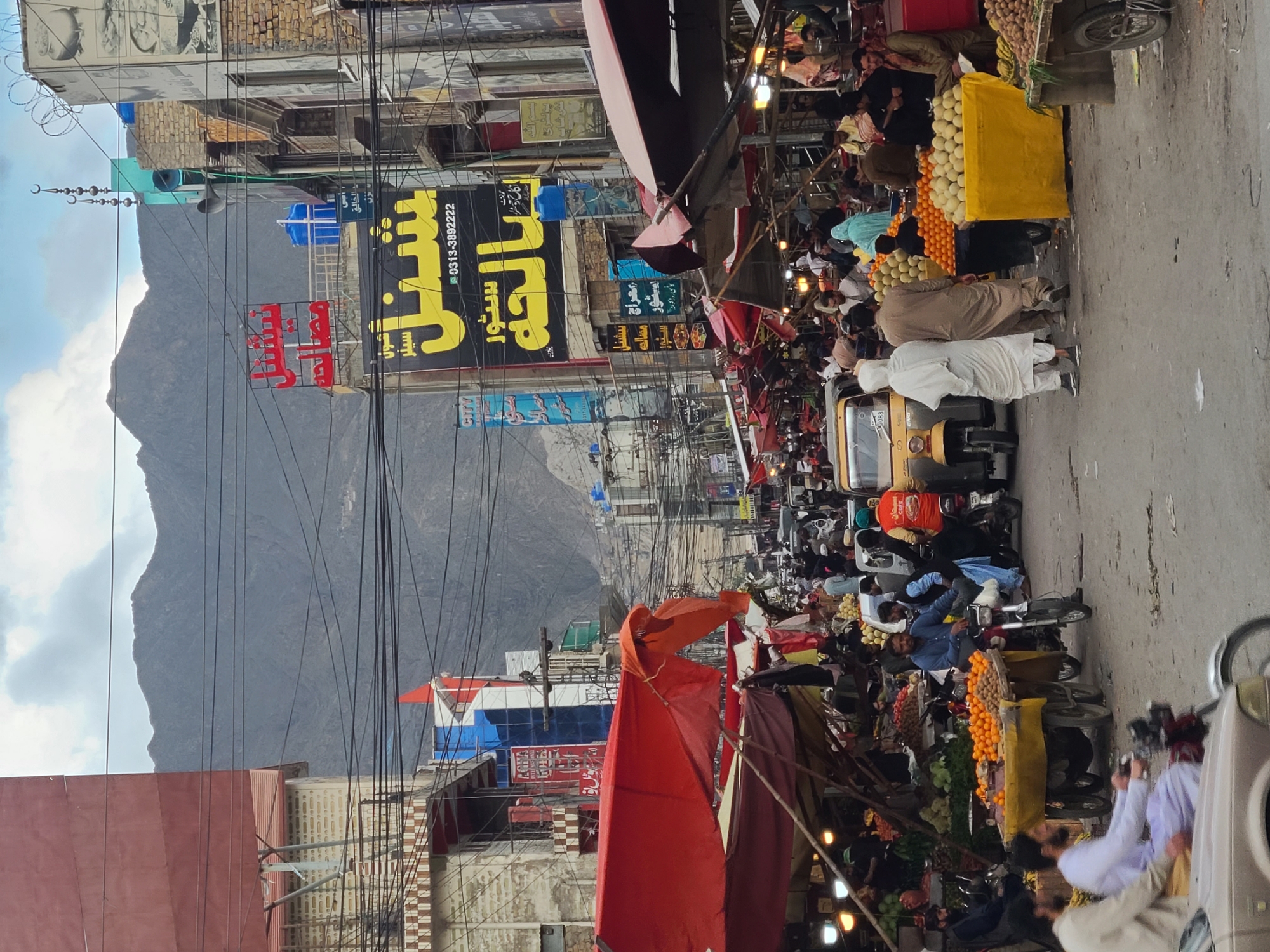 This image depicts a densely populated street market in Quetta City Tehsil, Pakistan, characterized by a bustling urban environment with a mountainous backdrop. The scene is observed from a slightly elevated perspective, looking down a street crowded with individuals, market stalls, and vehicles.
The foreground features a partial view of a silver-colored vehicle on the bottom left. The street surface appears to be a mix of concrete and dirt. In the mid-ground, numerous people, predominantly male, are present, engaging in various activities such as walking, standing, and operating vehicles. Many are dressed in traditional South Asian attire, including shalwar kameez. Several three-wheeled carts, heavily laden with produce like oranges and melons, are parked along the street. One cart is covered with a yellow tarp, displaying a pile of yellowish oval fruits, possibly potatoes or another variety of melon. Another cart is piled high with oranges. Multiple vendors are situated behind or near these stalls, interacting with potential customers. Motorcycles are visible, with some being ridden and others parked. A yellow auto-rickshaw is identifiable on the left side of the street.
Market stalls line both sides of the thoroughfare, featuring temporary awnings and tarpaulins, primarily in shades of red and orange, creating shaded areas. Underneath these coverings, various goods are displayed, illuminated by small artificial lights in some sections.
The background consists of multi-story buildings, appearing constructed from a mix of brick and concrete, some with visible wear. Overhead, a dense network of black electrical wires crisscrosses the sky. Numerous commercial signs in Arabic/Urdu script are mounted on the buildings. A prominent black sign with yellow text displays "شمل سٹور الحجہ" and a phone number "0313-3892222". A red sign above it reads "نیشنل مصالحہ". On the right, a building features a turquoise structure with multiple loudspeakers and a crescent moon and stars finial, indicative of a mosque or religious institution. Barbed wire is visible on nearby rooftops. Large, rugged mountains, dark in color, dominate the distant horizon under a partly cloudy sky.