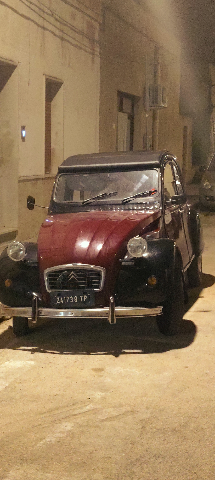 An aged, dark red and black Citroen 2CV car, with license plate "241738 TP", is parked on a textured, sandy ground in what appears to be a narrow street or alleyway in the town of San Vito Lo Capo, Italy. The car is facing the camera and is illuminated by artificial light sources, creating distinct shadows. The background consists of a light-colored building with an open doorway and an air conditioning unit mounted on the exterior wall. The scene is set at night.