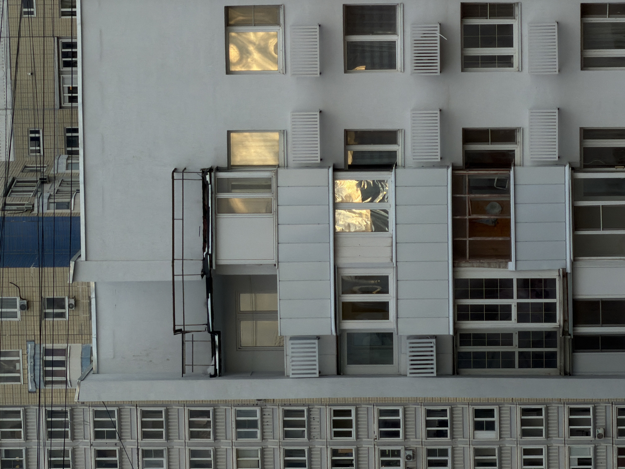 The image captures a high-angle, detailed view of the facades of two adjacent buildings, or sections of a single larger structure, located in Khimki, Russia. The perspective emphasizes geometric patterns created by windows and architectural elements.
The prominent building occupying the right and center of the frame features a light grey, almost off-white, facade. Its windows vary in size and design; many display strong, golden-yellow reflections, indicative of sunlight at a low angle, suggesting either early morning or late afternoon. Some windows are partially open, revealing darker interiors, while others are accompanied by white, horizontal slat-style blinds or shutters. Below a pronounced horizontal ledge, a grid of smaller, uniform rectangular windows with white frames extends towards the bottom of the image. White horizontal cladding panels are also visible on various parts of this building's exterior. A dark metal structure, possibly a ladder or utility frame, is affixed to the left side of this main facade.
To the left, a contrasting building section with a darker, possibly brick or concrete, facade is visible. It features smaller, regularly spaced windows with dark frames, some also reflecting light. A section of a blue awning or roof is present on this structure. Numerous dark cables crisscross vertically and diagonally across the left portion of the image, partially obscuring the darker building.
No human activity is depicted; the scene is a static architectural observation. The clear, bright reflections suggest a sunny day. The utilitarian architectural style and the visible external cabling are consistent with urban environments in the specified location of Khimki, Russia. There is no visible text or signage in the image. The overall composition highlights urban density and repetitive structural elements.