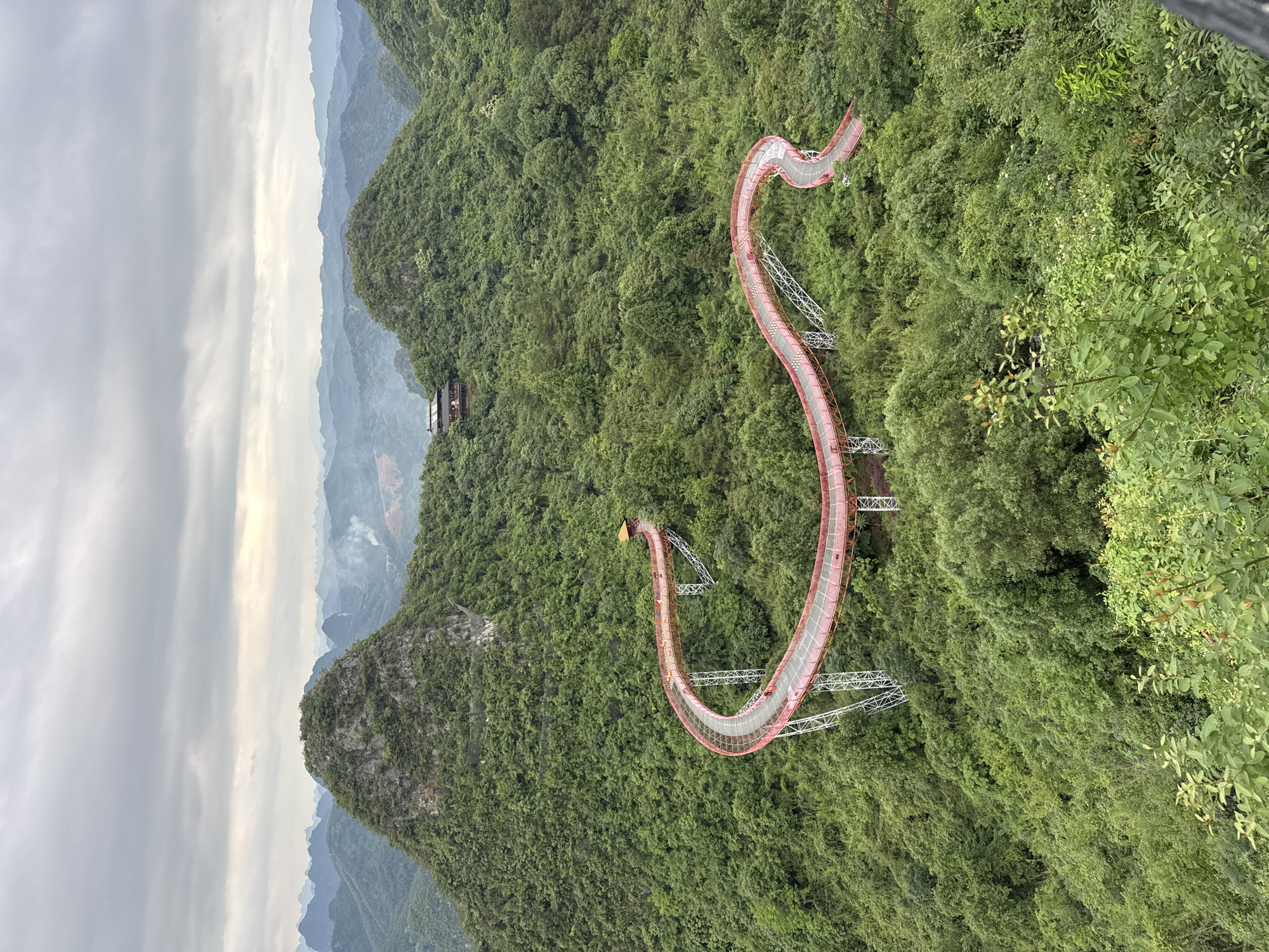 The image depicts a high-angle view of a walkway winding through a densely forested mountainous area. The walkway is predominantly red, supported by white, cross-braced support structures, and it snakes along the contour of the landscape. Lush green trees cover the surrounding terrain, which is characterized by steep hills and valleys. In the background, additional mountain ranges are visible under an overcast sky.
The scene is of the Guilin Yulong River Bamboo Raft tourist area in Guilin, China.
