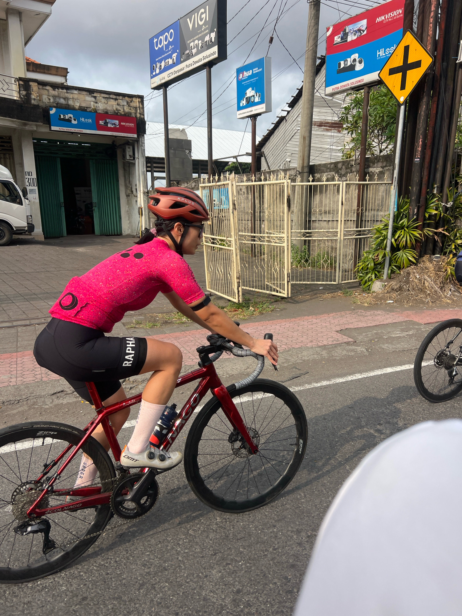 In Denpasar, Indonesia, a female cyclist in a bright pink jersey and black cycling shorts is riding a red road bicycle on a paved road. She wears a reddish-brown helmet and white cycling shoes. The street features a red brick pedestrian path and a metal fence along the side. In the background, several billboards advertise security camera brands like "Vigi," "Tapo," "Hikvision," and "Hilook." A white delivery truck is parked to the left. The sky is overcast.