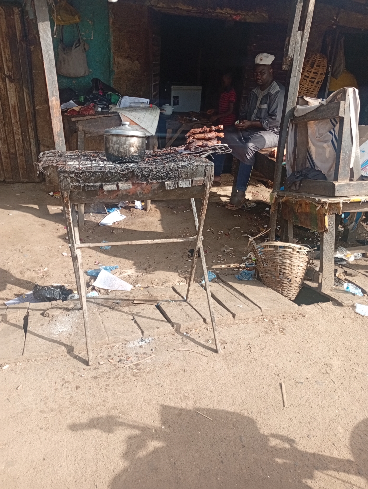 The image depicts an outdoor scene, likely a market stall, in Rafin Par, Nigeria. In the foreground, a charcoal grill, blackened and covered with what appears to be burnt food debris, sits on a sandy, uneven surface. Scattered around the grill are various pieces of trash, including plastic bags and paper.
Behind the grill, a man wearing a grey long-sleeved shirt, dark pants, and a white cap is seated on a wooden bench. He is holding some items, possibly money, in his hands. To his left, a younger person in a red and black striped shirt stands behind a counter or table, seemingly engaged in serving or preparing food. A pile of cooked meat, likely grilled, is visible on a plate or surface in front of this person.
The background reveals a makeshift structure made of wood and corrugated metal, suggesting a temporary or semi-permanent stall. Bags and other items are hung on the walls or structures. A woven basket is placed on the ground to the right of the frame, near the seated man. The overall environment appears rustic and somewhat unkempt, typical of many market settings in developing urban areas. The lighting indicates a bright, sunny day.