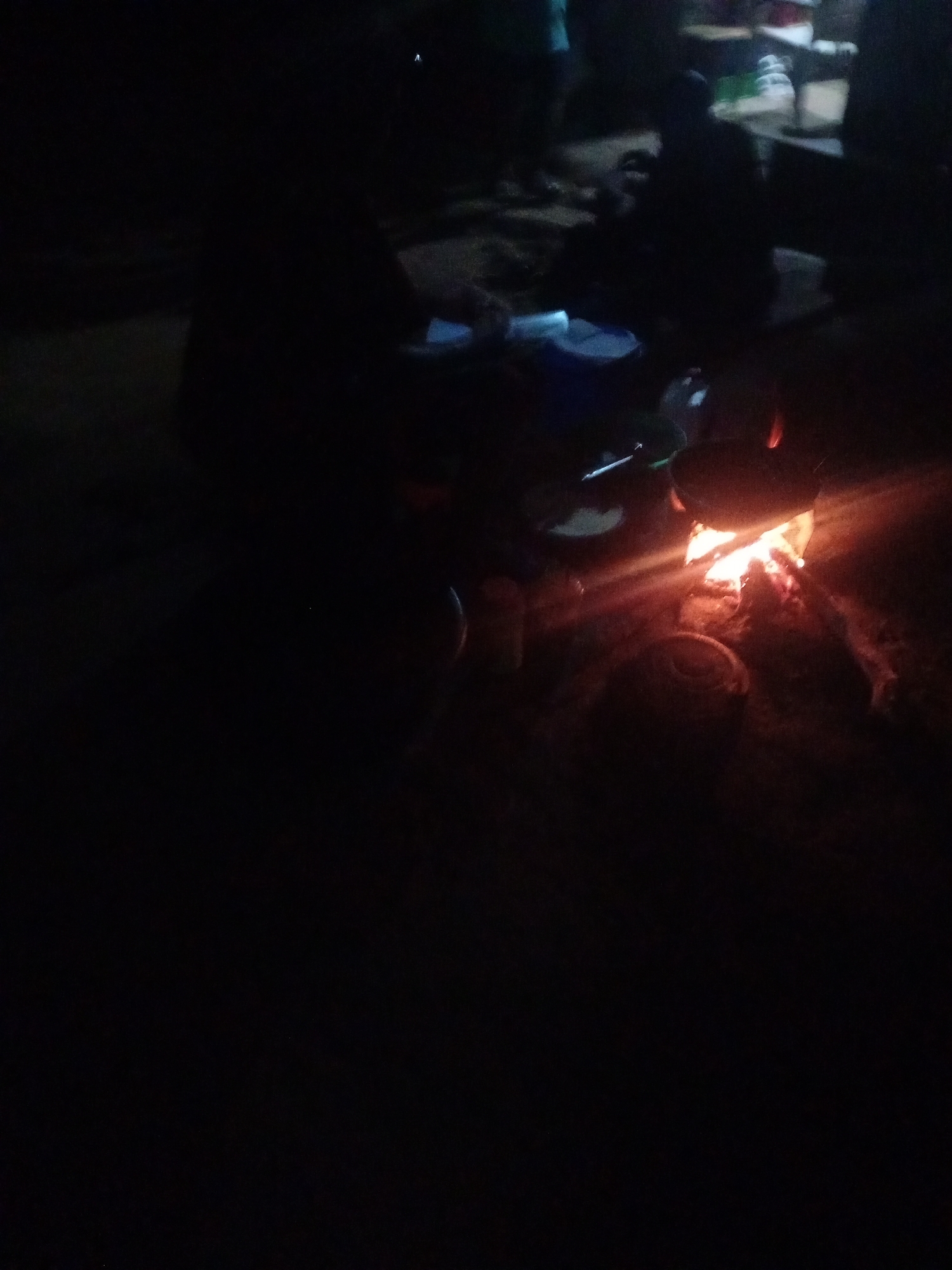 In the dim light of a campfire, several individuals are gathered in what appears to be an outdoor setting in Sabon Gari, Nassarawa, Nigeria. The scene is primarily lit by the orange glow of the fire, casting long shadows and obscuring many details.
The subjects appear to be a small group of people, mostly silhouettes against the darkness. Their exact age or gender is difficult to discern. There are objects that suggest cooking or food preparation around the fire. Several metal pots or containers are visible near the flames, some of which may contain food. A long metal rod, possibly a cooking utensil or part of the fire structure, is also present. The ground around the fire is rough and appears to be dirt or sand.
The activity taking place is a communal gathering around a campfire, likely for cooking or socializing. The time of day is clearly night, indicated by the darkness and the reliance on firelight. There are no obvious cues about the weather, but the nature of the gathering suggests it is not raining. The location is identified as Sabon Gari, Nassarawa, Nigeria, suggesting a rural or semi-rural community setting. The overall mood is one of shared activity, with the warmth of the fire providing comfort in the darkness. No text is visible in the image.