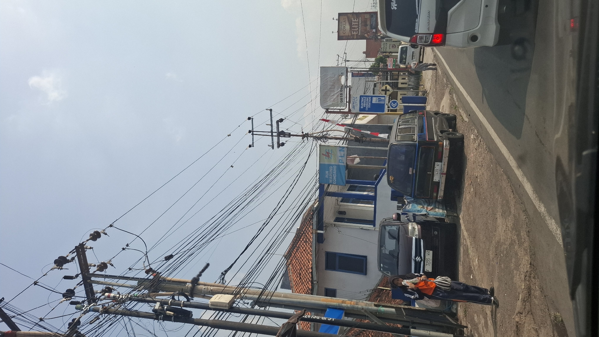 The image captures a street scene in Cikarang, Indonesia, characterized by a dense network of overhead power lines. On the left, a building with a white facade and a red-tiled roof appears to be a PLN (Perusahaan Listrik Negara) office, indicated by signage. Parked in front of the building are two vehicles: a dark-colored MPV and a black van. A young woman in an orange vest and headscarf stands on the side of the road, holding food. The street itself is paved, with a white lane marker visible. Further down the road, more vehicles, including a white SUV, are present, suggesting moderate traffic. Several billboards are also visible along the roadside, advertising various products and services. The sky is clear with scattered clouds.