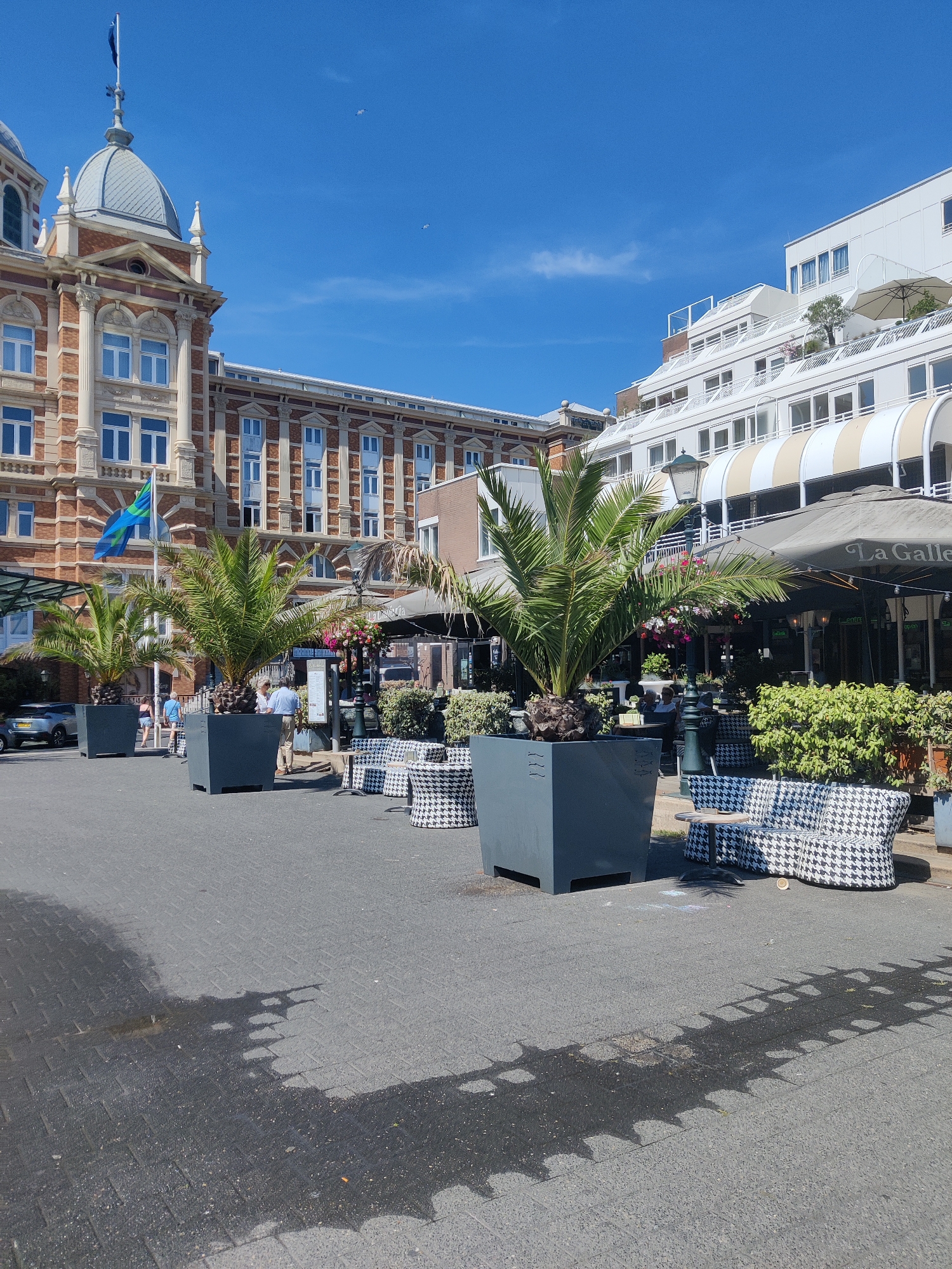 The image depicts a street scene in The Hague, Netherlands, likely in a commercial or recreational area. In the foreground, a paved walkway is visible, with a noticeable wet patch that has cast a shadow of a fence-like pattern onto the ground. Several large, dark grey planters containing palm trees are strategically placed on the walkway. Adjacent to these planters are outdoor seating areas, featuring sofas and chairs upholstered in a black and white houndstooth pattern, with small circular tables.
In the background, a multi-story building with an ornate brick facade and a domed roof dominates the left side of the frame. A flag with blue and green stripes flies from a flagpole in front of this building. To the right of this building, a more modern, white multi-story structure is visible, characterized by a series of balconies and awnings. Below these buildings, an outdoor seating area of a restaurant or cafe is set up, with tables and chairs under a large grey umbrella that bears the name "La Galler". Green bushes and some potted plants add to the ambiance of the outdoor dining space.
Several individuals are present in the scene, mostly appearing as patrons or staff of the establishment. Two individuals are seen standing near a menu board outside the cafe. Another individual is seen walking away from the camera on the left side of the walkway. A silver car is parked on the left side of the image. The sky is a clear, vibrant blue with a few wispy clouds and two small white birds in flight. The overall impression is a sunny day in an urban environment, likely a promenade or a square adjacent to businesses and establishments.