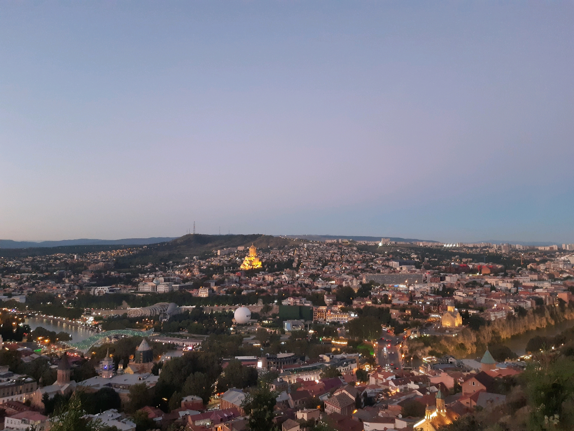 This is a panoramic, high-angle, dusk view of Tbilisi, Georgia. The scene overlooks a dense urban landscape with numerous buildings, illuminated by streetlights and building lights. In the mid-ground, a river, likely the Kura River, is visible, with bridges and lights along its banks. Prominent structures include the Holy Trinity Cathedral of Tbilisi, illuminated gold on a hilltop in the distance, and several other churches with distinctive domes. Also visible is a large, white, spherical building. The city is nestled within rolling hills, with the sky transitioning from a pale blue to a darker hue as twilight deepens. No people are discernible in the image.