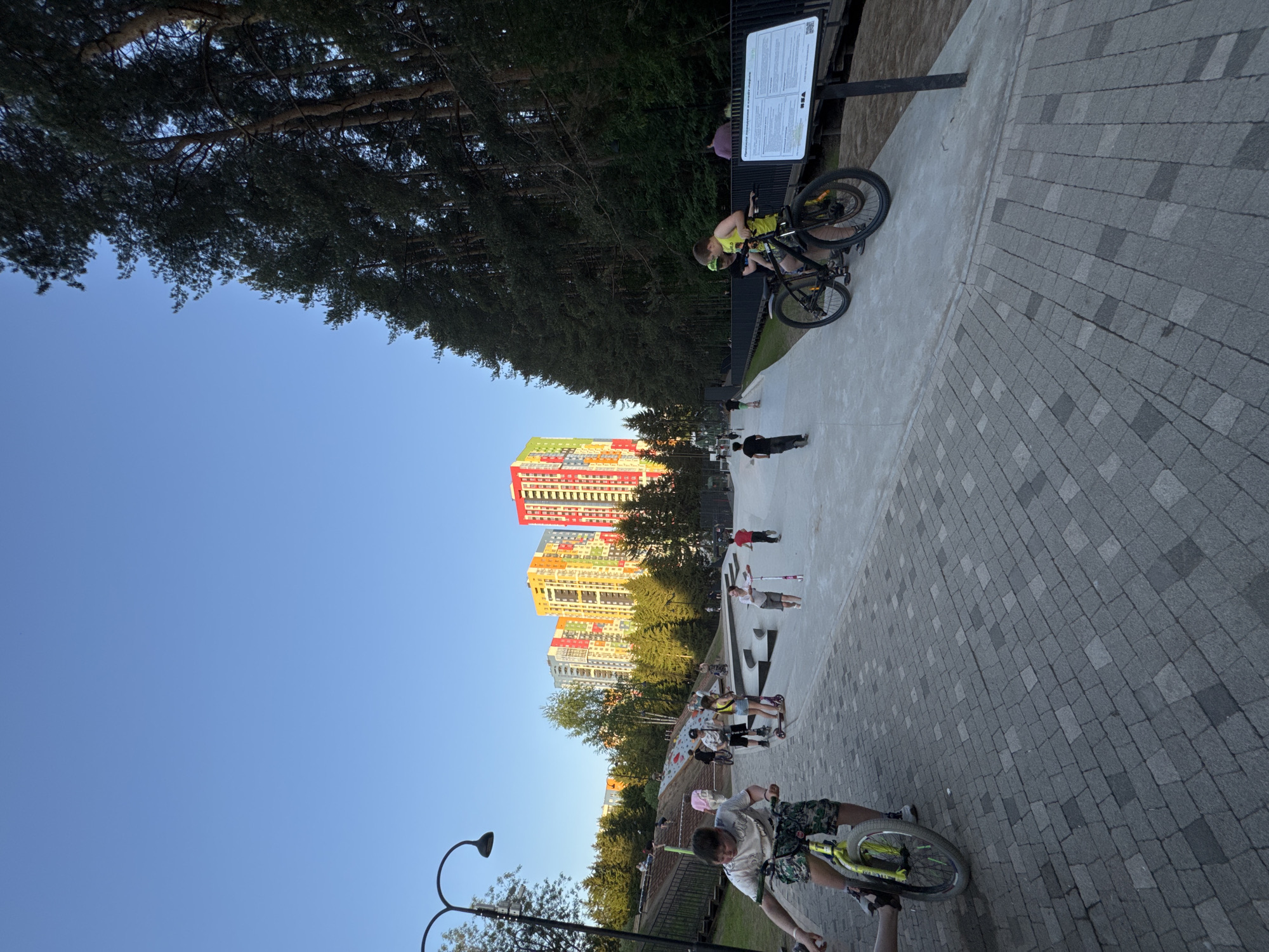 The image captures a skate park in Izhevsk, Russia, situated adjacent to brightly colored residential buildings. The ground is paved with grey brick. Multiple people are present in the park, including two individuals with bicycles at the front and top of the image, as well as several people with scooters and skateboards. A signpost is seen behind one cyclist. The sky is clear and blue, while trees border the park.