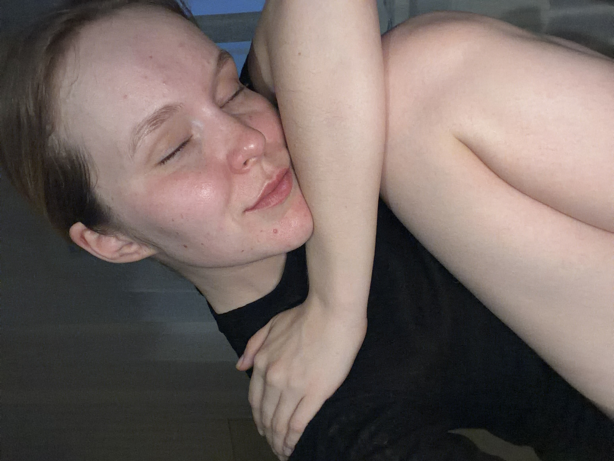 A close-up captures a fair-skinned person with closed eyes, resting their cheek against a bare arm and shoulder. The person has short, light brown hair and a visible ear. Their skin exhibits freckles and minor blemishes. They are wearing a black shirt. The background shows a hint of a window. The location is not discernible from the image. There is nothing in the image to indicate the location is Izhevsk, Russia.