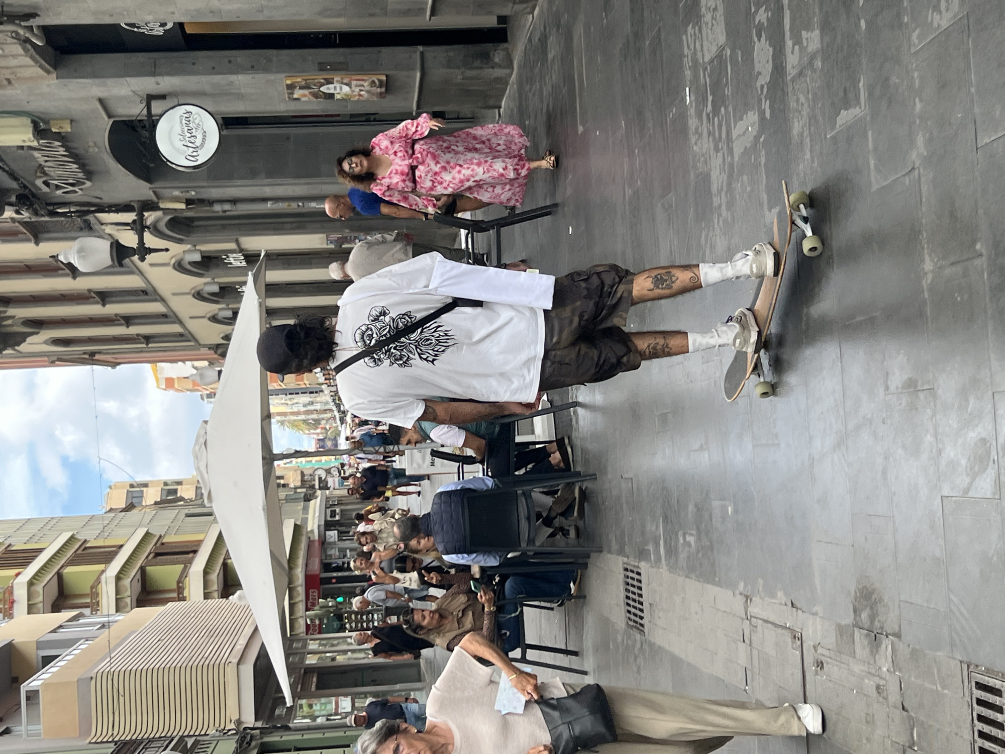 The image captures a street scene in Las Palmas De Gran Canaria, Spain. The location appears to be a pedestrian area lined with buildings. Several people are present, some seated at outdoor tables under a large white awning, likely part of a cafe or restaurant. One person is standing on a skateboard, and another is seated nearby. One woman in a pink floral dress is seated at a table, while other people are walking by. The street surface is tiled with what appear to be light-grey slabs. The sky is partly visible. There are streetlights and hanging signs along the buildings. The scene is taken at street level.