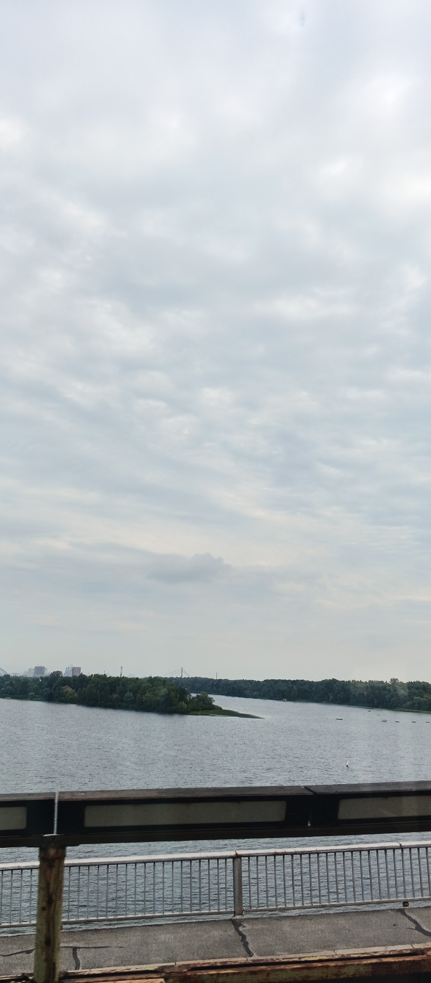 The image depicts a serene, overcast day over a wide body of water, likely a river or reservoir, in Kyiv, Ukraine. In the foreground, a metal railing of a bridge or overpass is visible, with a weathered concrete walkway. Beyond the railing, the water stretches out, reflecting the muted tones of the sky. A small, densely wooded island juts out into the water on the left side, with buildings and structures visible on the distant shoreline behind it. Further to the right, a long stretch of forested shoreline extends into the distance, with a thin bridge or structure faintly visible on the horizon. Several small, dark objects, possibly buoys or small boats, are scattered across the water. The overall atmosphere is calm and natural, with a hint of urban presence in the background.