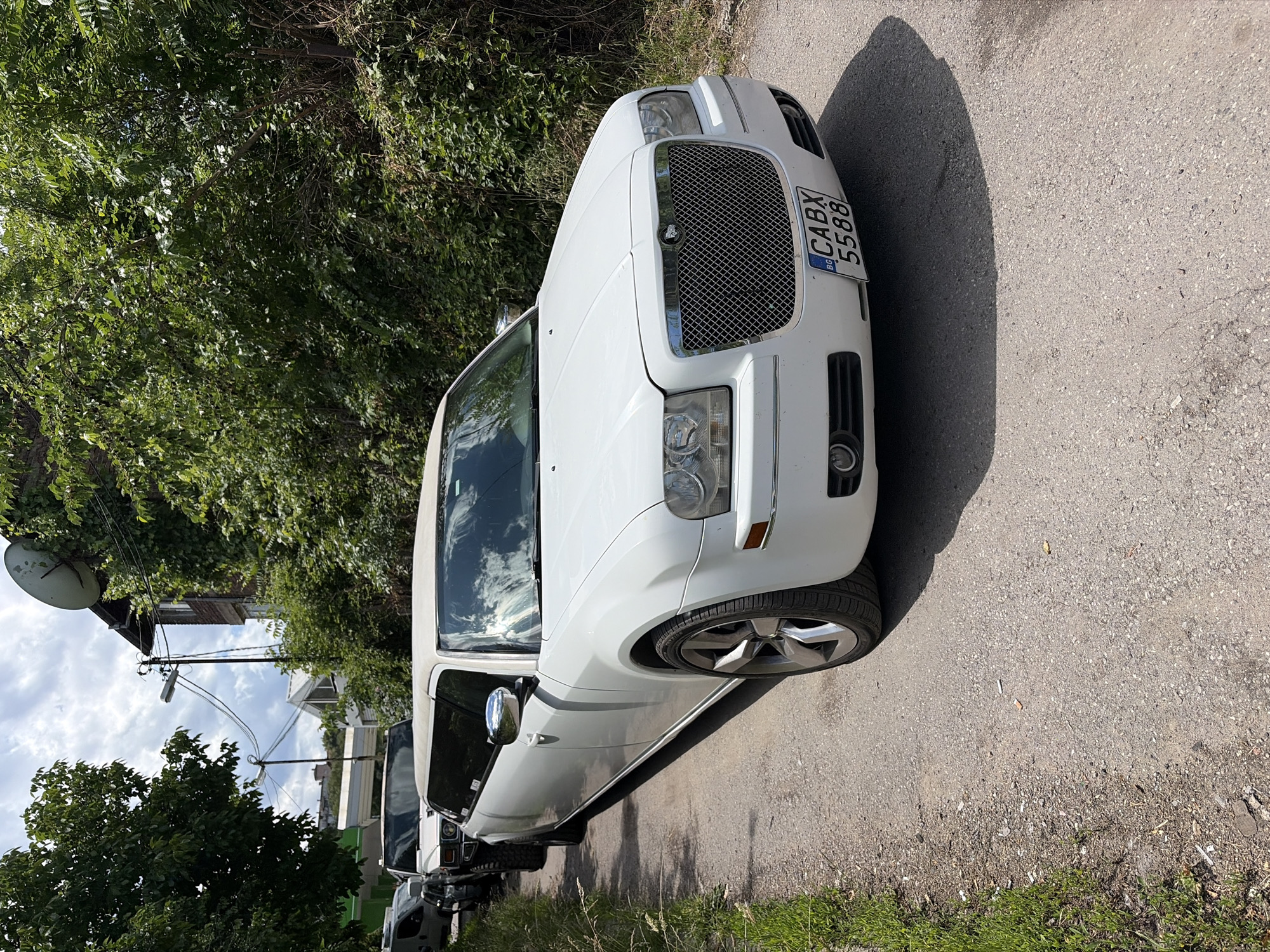 A white Chrysler 300 stretched limousine is parked on a narrow, unpaved road in Sofia, Bulgaria. The limousine is the primary subject, occupying the foreground and right side of the image. Its front end, including the grille, headlights, and license plate (BG CABX 5588), is clearly visible. The limousine is flanked by lush green trees and bushes on the right and a grassy verge on the left. Behind the limousine, other vehicles, including a white Hummer H2 limousine and a white van, are also parked along the road, suggesting a convoy or a gathering. In the background, a building with a satellite dish on its roof and utility poles with overhead wires are visible, indicating an urban or semi-urban environment. The sky is partly cloudy. The scene appears to be a daytime shot.