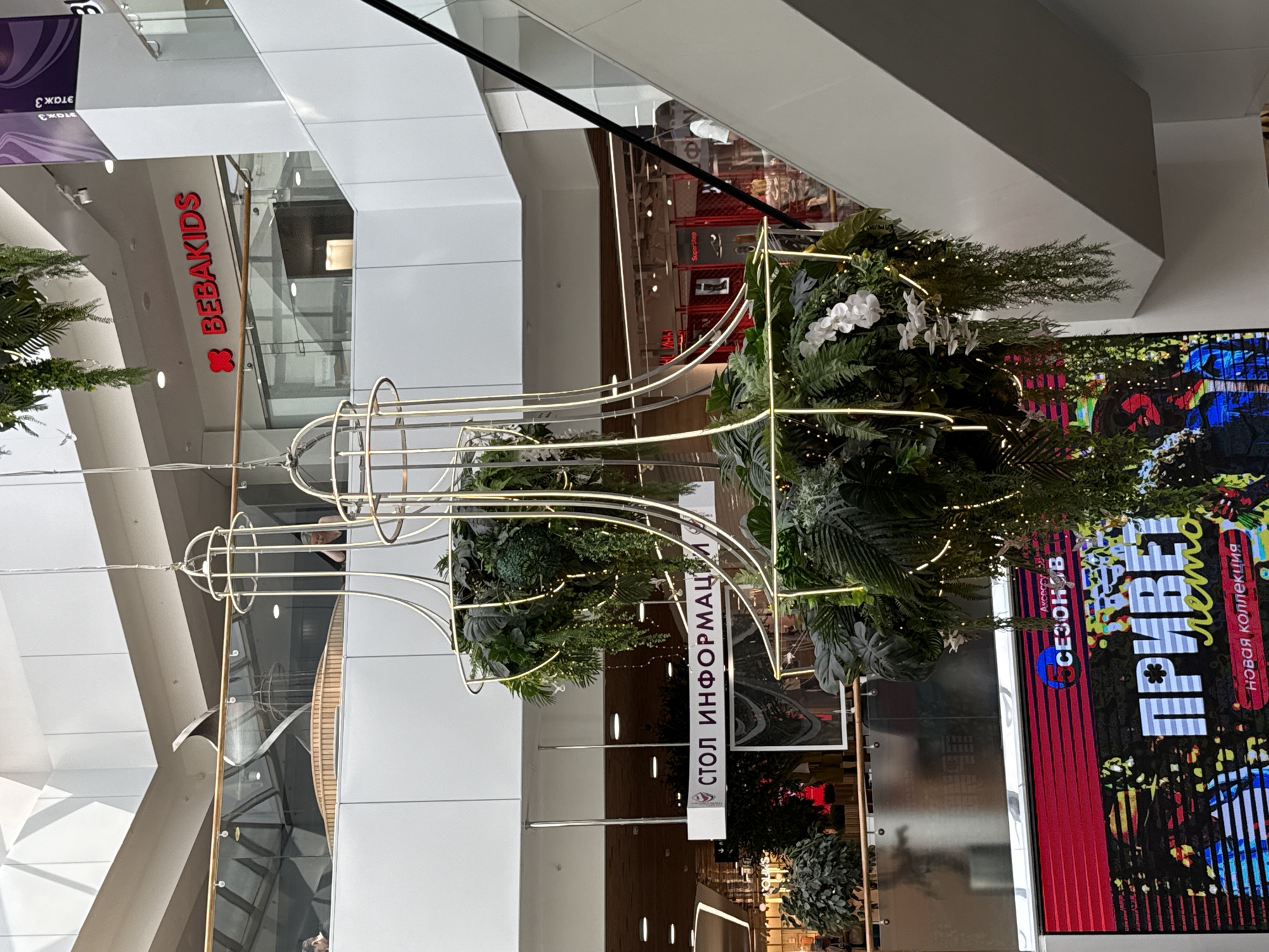 The image depicts the interior of a shopping mall, likely in Izhevsk, Russia, given the text visible on signage. In the foreground, a large, ornate hanging decoration featuring artificial plants, white orchids, and string lights is prominently displayed. The decoration is suspended from the ceiling by thin wires, creating a chandelier-like effect.
Behind the hanging decoration, a sign with Russian text reads "СТОЛ ИНФОРМАЦИИ," which translates to "Information Desk." Further back, a store sign with red lettering and a stylized red bow reads "BEBAKIDS." Another sign above this store appears to have Cyrillic text related to floor level, possibly indicating "Floor 3."
To the right of the frame, a white escalator is visible, with its black handrail curving upwards. A person in a white shirt can be seen on the escalator. In the middle ground, a digital screen displays a colorful advertisement with prominent Russian text, including "ПРИВЕТ лето," meaning "Hello summer," and "новая коллекция," meaning "new collection." A circular logo with the text "СЕЗОНВ" is also present on the screen.
The mall features multiple levels with white railings and balconies, and the ceiling is a multi-faceted white structure with angled panels and visible lights. The overall environment suggests a modern, well-lit commercial space designed for shopping.