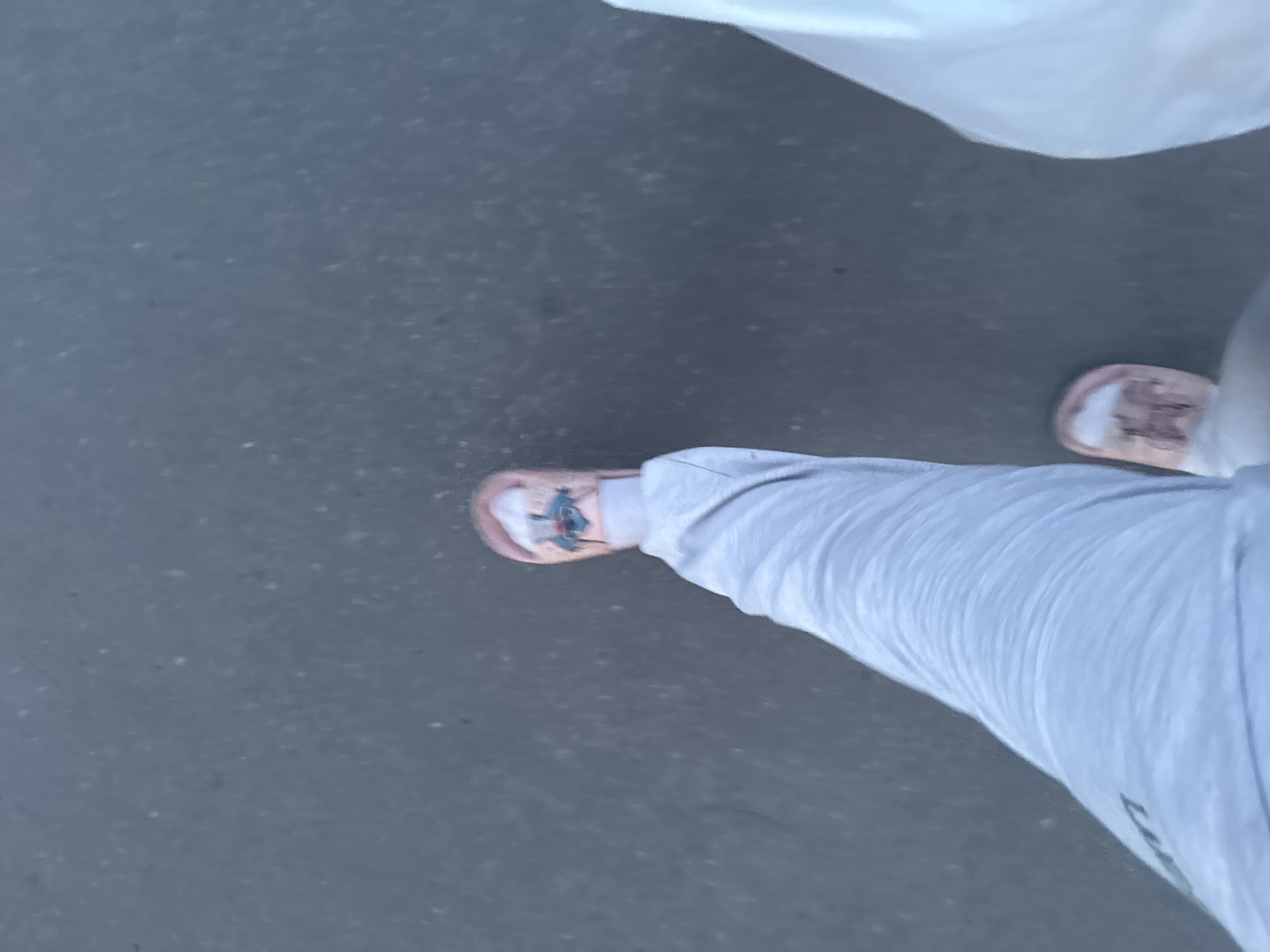 This is a first-person perspective image taken outdoors, likely on a paved surface such as asphalt, in Гожули, Ukraine. The focus is on the lower legs and feet of the individual. The person is wearing light grey sweatpants and light pink slides with a cartoonish graphic on the right slide. The left leg is bent at the knee, with the foot slightly forward. To the right of the frame, a portion of a white, opaque plastic bag is visible. The ground appears textured with small specks, consistent with asphalt. The lighting suggests it is daytime, possibly late afternoon due to the shadows.