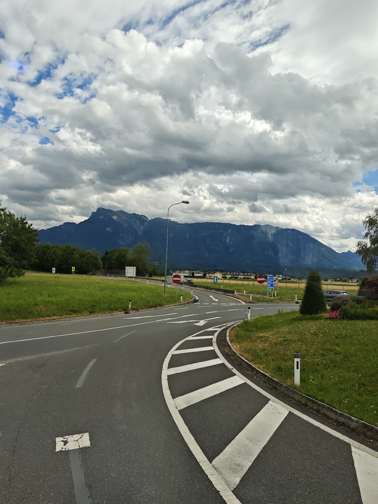 This media file captures a scenic view of a road curving through a grassy landscape with a majestic mountain range in the background. The scene appears to be set in Walserfeld, Austria.
The foreground features a paved road with clear white lane markings, indicating a junction or an approach to one. The road curves to the right, with a grassy embankment and a landscaped area to its side. Several white bollards line the edge of the road, and there are also some small white rectangular markers on the road surface.
In the middle ground, a lush green field stretches out, bordered by trees. Beyond the field, a line of buildings can be seen, suggesting a small town or village. Traffic signs are visible along the road, including a red and white circular sign indicating "no entry" and a blue sign with white text, likely providing directional information.
The background is dominated by a large, dark mountain range, its peaks and slopes shrouded in a mix of sunlight and shadow. The sky above is a dynamic canvas of dramatic clouds, with patches of blue sky peeking through. Some of the clouds are heavy and dark, suggesting potential rain, while others are bright and white, illuminated by the sun.
A solitary street lamp stands on the right side of the road, its light directed downwards. There are no people visible in the image. The overall impression is one of natural beauty and open space, with the road serving as a pathway through the landscape. The weather appears to be changeable, with a mix of sunshine and heavy cloud cover, typical of a summer day in a mountainous region. The time of day is likely afternoon, given the lighting conditions. The location cues, such as the road layout and the specific appearance of the mountains, are consistent with a European setting, specifically Austria as indicated by the location context.