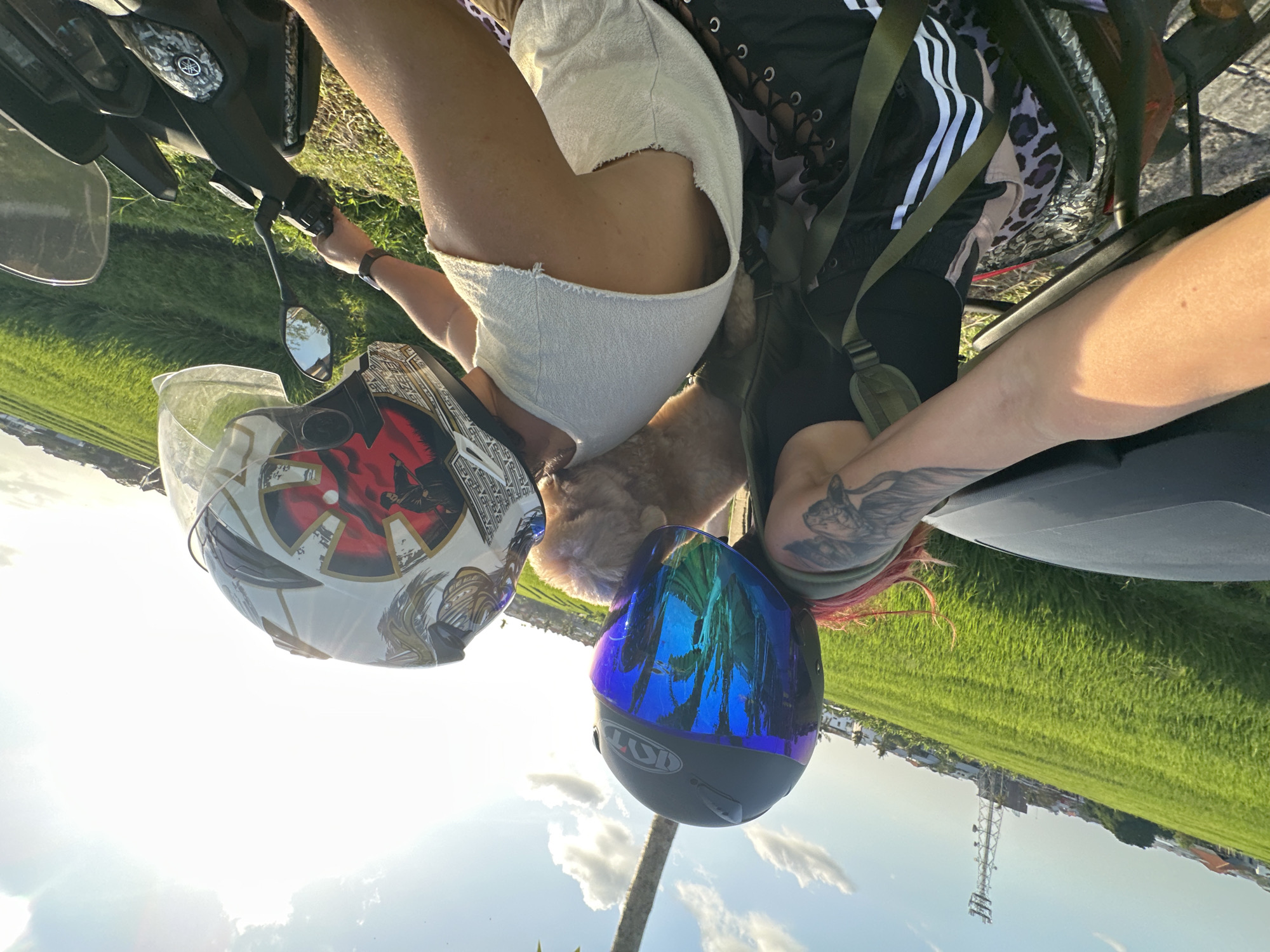 Two individuals wearing helmets are riding a scooter through a lush green rice field with a clear blue sky and scattered clouds. The person in the foreground, on the left, has red hair and is wearing a black tank top with a tattoo on their arm. They are holding a small, light-brown dog on their shoulder. The person in the back, on the right, is wearing a white sleeveless shirt and a helmet with a graphic design. The scooter is black and has a Yamaha logo on the front panel. In the background, there are buildings and a cell tower, suggesting an urban or semi-urban environment. The image appears to have been taken in North Kuta, Indonesia, given the landscape and architecture.