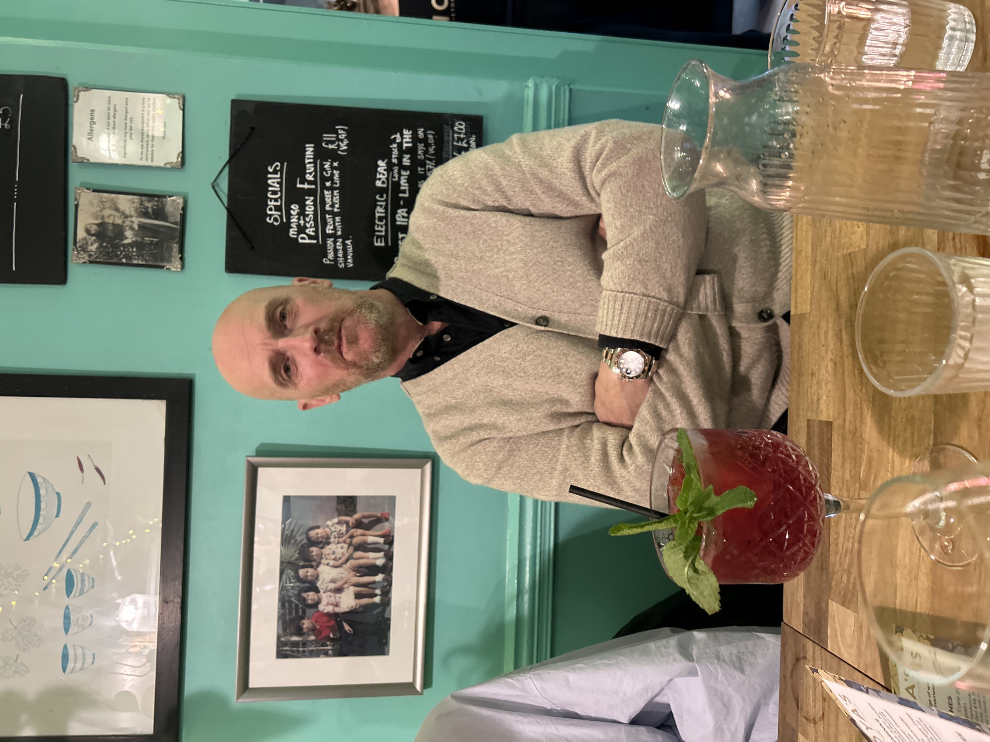 The image shows the interior of a restaurant or bar, possibly in Bath, United Kingdom, based on the presence of signs and the overall atmosphere. A man with a shaved head, wearing a beige cardigan over a dark shirt and a watch, is seated at a table with his arms crossed. A red cocktail in a patterned glass, garnished with mint and a black straw, sits on the table next to empty glassware, including a carafe. The table is wooden.
Behind the man is a light turquoise wall adorned with framed artwork and blackboards. The blackboards display the word "SPECIALS" and menu items, including a Mango Passion Fruitini and Electric Bear IPA. The wall also displays allergy information and other framed images. The overall environment suggests a casual dining or drinking establishment.