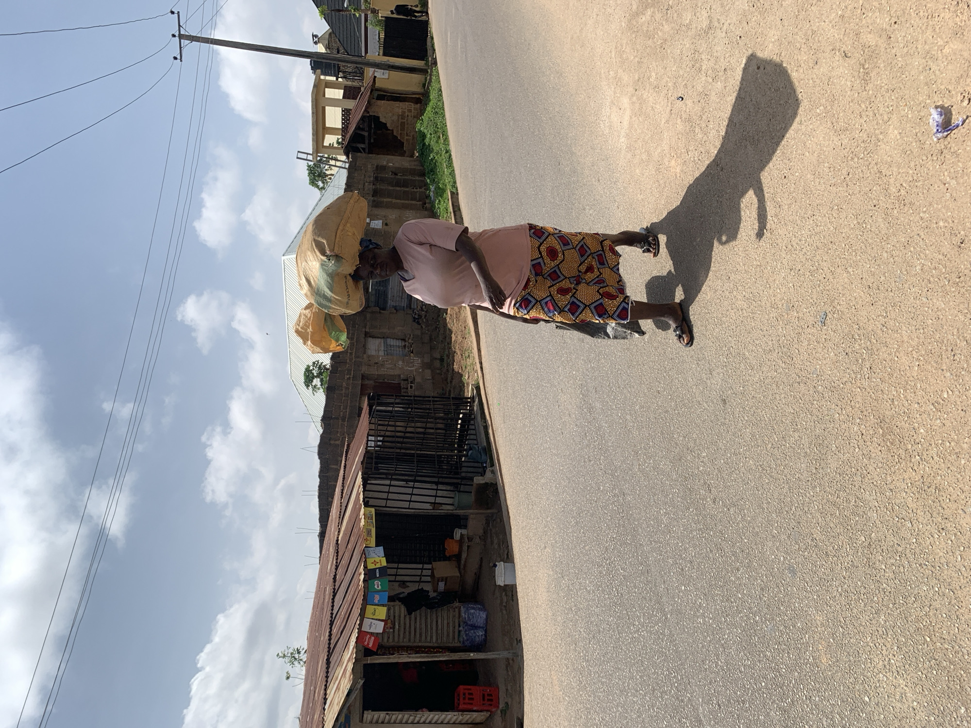 A full-body, eye-level shot captures a woman walking on an asphalt road in Jos, Nigeria, carrying two large sacks of goods balanced on her head. She is wearing a light pink t-shirt and a colorful patterned skirt. Her feet are adorned with brown sandals. To the left of the road, a small roadside shop with a corrugated metal roof displays various items, including crates of drinks and bagged goods. The shop is secured with metal bars on its windows and doorway. The background reveals a street with buildings and a power pole. The sky above is a mix of blue and scattered white clouds. The sun casts a long shadow of the woman on the road to her right.
