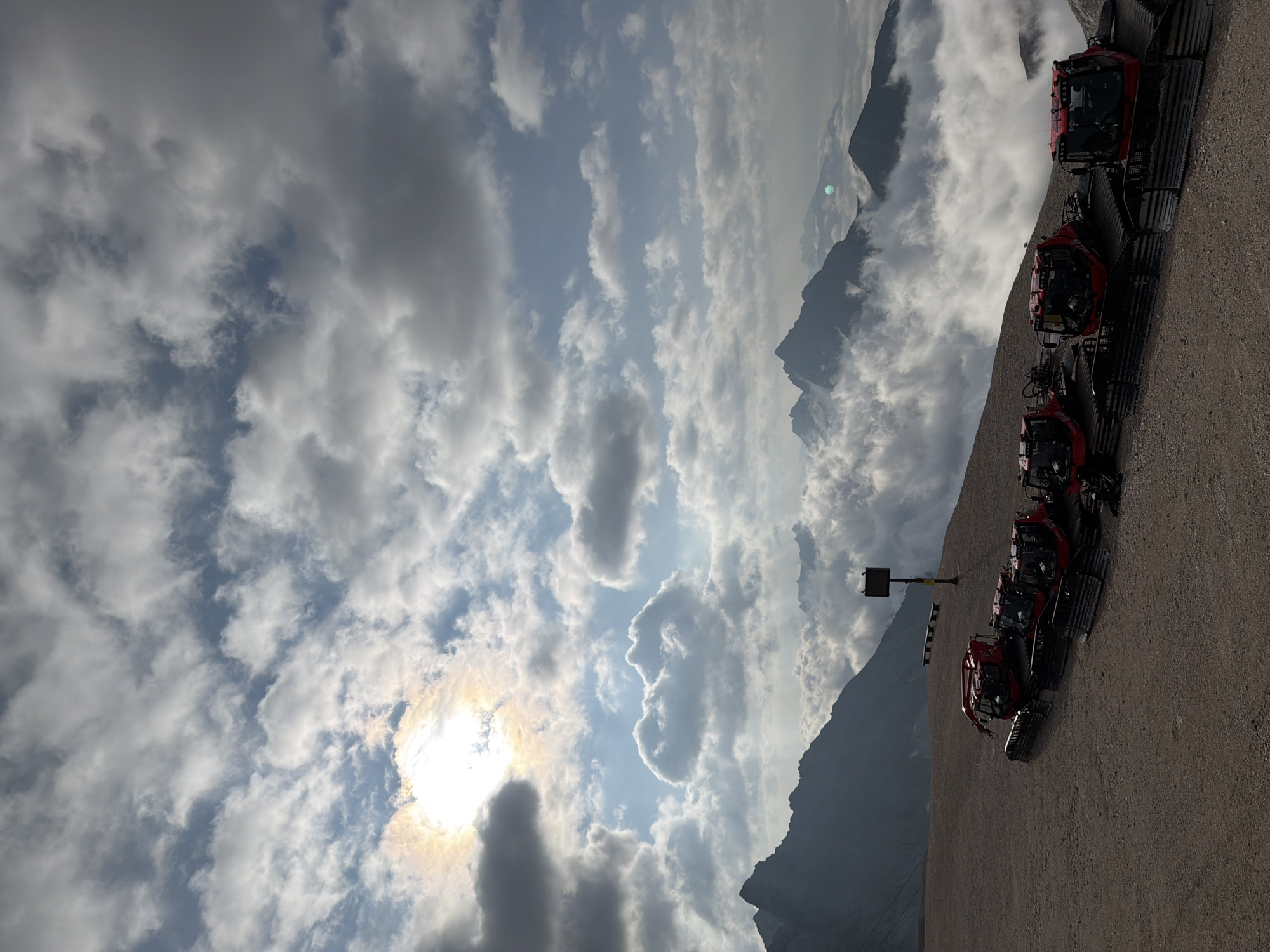 The image depicts a view from a mountain summit overlooking a cloud-filled valley with snow groomers parked in the foreground. The sun is visible in the upper left portion of the sky, partially obscured by clouds, casting a bright light. Several red snow groomers are lined up on a gravel surface. In the background, jagged mountain peaks rise above the clouds, with some appearing to be in shadow. The overall scene suggests a high-altitude location, possibly a ski resort, given the presence of the snow groomers. The location is identified as Garmisch-Partenkirchen, Germany, specifically at the Zugspitze, Germany's highest peak.