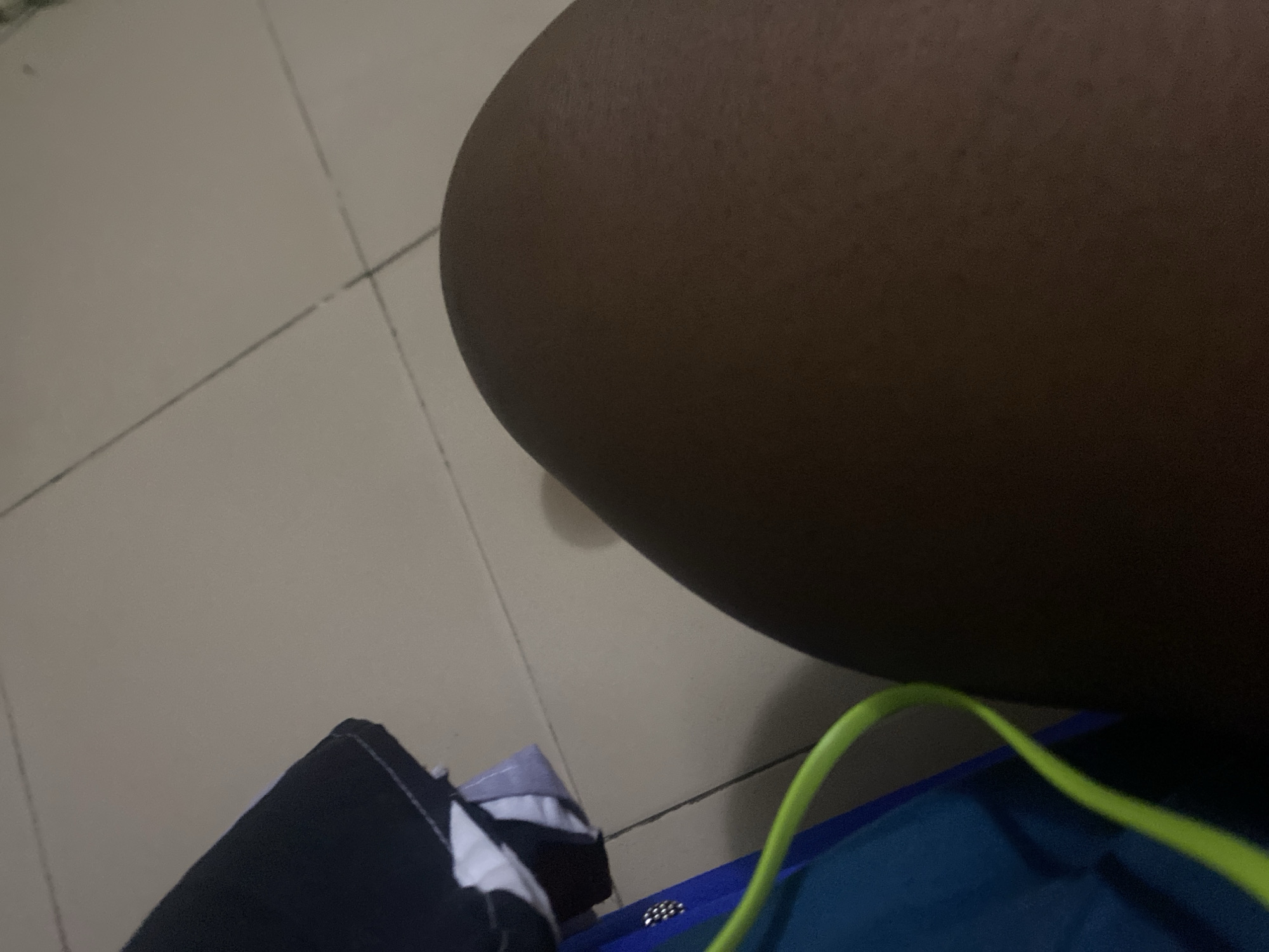 A close-up, overhead shot shows a person's bare leg and thigh in the foreground, appearing to be of African descent. The skin is a medium to dark brown. Behind the leg, on the left side of the frame, a blue bag with a lime green strap is visible. A black and white piece of clothing is draped over the bag. The background consists of a light-colored tiled floor with visible grout lines. The scene is lit by artificial light, casting a shadow of the leg onto the floor. There are no discernible city or country indicators in the image; therefore, its location in Howayi, Nigeria cannot be confirmed.