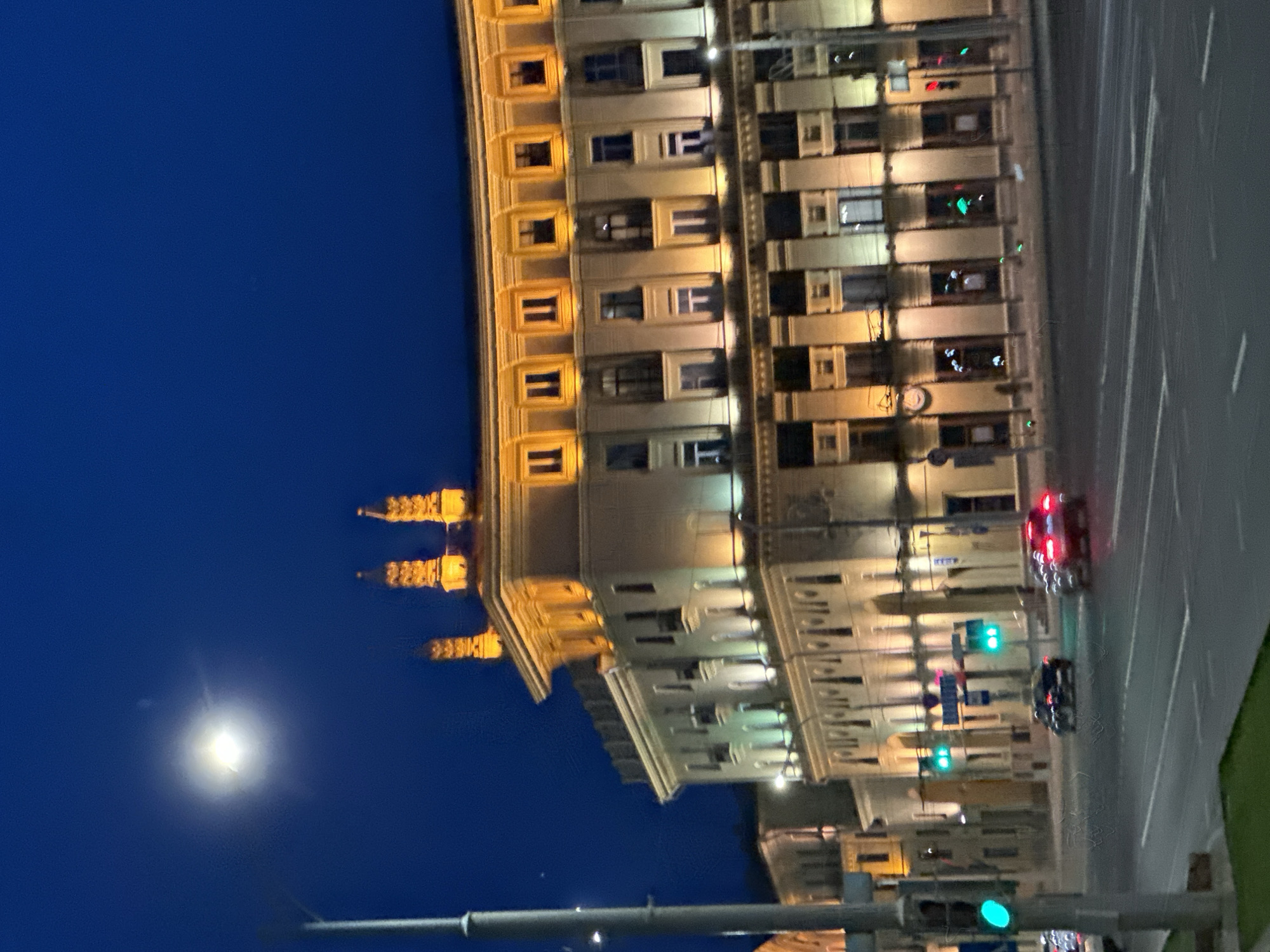 The image depicts a nighttime urban scene focusing on a prominent illuminated building and a street with vehicular traffic. **Environment:** The setting is an urban area at night, under a clear, deep blue sky. A very bright, overexposed spherical light source, consistent with the moon, is visible in the upper left quadrant. The air appears clear. **Location:** The main building in the image is identifiable as the **Central Post Office** (Main Post Office) located at Nezavisimosti Avenue, 10, in **Minsk, Belarus**. **Subjects and Objects:** * **Building:** A large, multi-story building, the Central Post Office, occupies the right and central portions of the frame. It features a classical architectural style with a cream or light-colored facade. The building is extensively illuminated: the upper cornice and series of rectangular windows below it glow with warm yellow light, as do three distinct, ornate spire-like structures on the rooftop. The lower floors are illuminated with a combination of warm and cooler white lights, highlighting window frames and decorative elements. The building follows a gentle curve along the street. * **Roadway:** A multi-lane asphalt road, Nezavisimosti Avenue, runs horizontally across the foreground and midground. It features visible white lane markings. * **Vehicles:** At least two passenger cars are present on the road in the midground, appearing to be in motion away from the viewer. One vehicle is dark-colored, and the other displays illuminated red taillights. A third vehicle is partially visible on the far left. * **Traffic Signals:** Multiple traffic lights are active. A tall grey pole on the far left foreground holds a green traffic signal facing the viewer. Further down the street, two additional green traffic signals are visible, one in front of the main building and another slightly to its right. A red traffic signal is visible on the far right, perpendicular to the main road flow. * **Street Furniture:** A prominent grey utility pole stands on the far left, extending from the bottom to the upper portion of the frame. Other smaller poles and blue rectangular street signs are visible in the midground. * **Vegetation:** A strip of green grass is visible in the immediate foreground, likely a median or sidewalk edge. * **Adjacent Structures:** A less illuminated building, also featuring historical architecture, is visible in the background to the left of the Central Post Office. **Actions and Interactions:** Two vehicles are observed in motion on the roadway, indicated by their