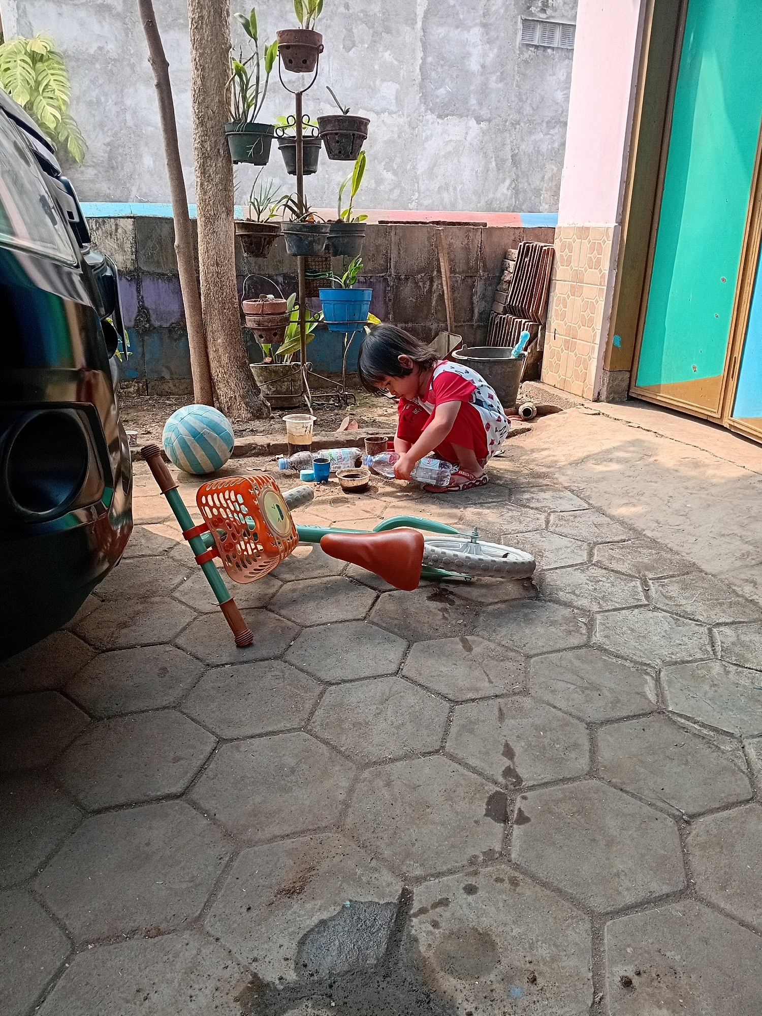 A young child, with dark hair and wearing a red and white patterned dress, is crouched on a paved outdoor area, playing with a plastic water bottle. The child is focused on the bottle, and their left hand is in the process of manipulating it. To the left of the child, a bicycle lies on its side, its orange basket and brown seat clearly visible. A blue and white striped ball is positioned behind the bicycle. The ground is covered with hexagonal paving stones. In the background, a black vehicle is parked. Further back, a wall with a colorful mosaic-like pattern is visible, along with several potted plants on stands. A large building with a pink facade and a bright turquoise door is to the right. The overall scene appears to be a residential outdoor space, possibly a yard or driveway, in Gebang, Indonesia.
