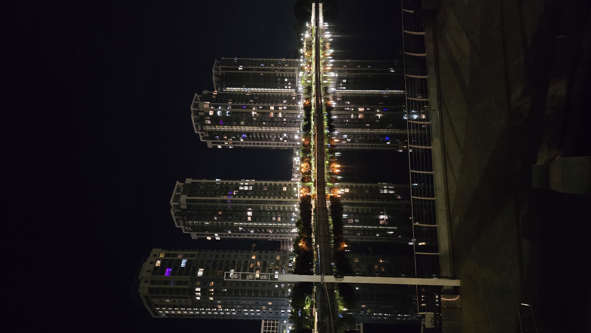 This image depicts a striking urban night scene, likely situated along a waterfront in Kyiv, Ukraine. The primary subjects are multiple modern, high-rise residential buildings, their facades dotted with numerous glowing windows. These windows exhibit a variety of light colors, including white, yellow, purple, and blue, suggesting occupied residences.
A long, narrow pedestrian pathway or bridge, illuminated by streetlights and bordered by trees or bushes, runs horizontally across the central part of the frame, separating the buildings from the dark, still water. The water acts as a mirror, perfectly reflecting the illuminated buildings, streetlights, and the architectural elements, creating a symmetrical and almost surreal effect.
On the far right of the image, a large, dark, and textured concrete wall or embankment is visible, alongside a vertical metal railing or fence structure. The overall setting is an urban environment at night, characterized by the stark contrast between the vibrant artificial lights and the deep black sky. The calm reflections in the water indicate still conditions, possibly suggesting fair weather. No people or specific events are visible, and the scene conveys a sense of peaceful urban solitude after dark. There is no visible text in the image.