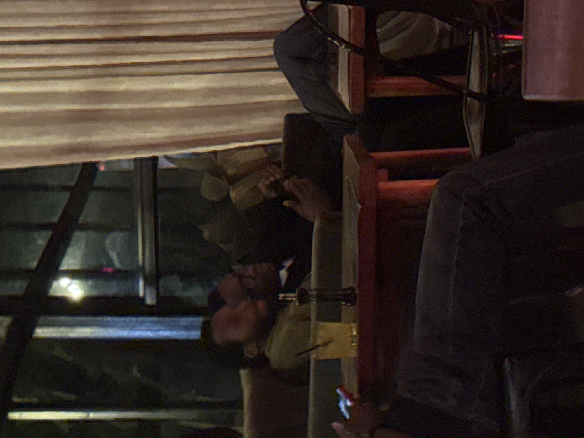 This image depicts an interior scene in the city of Abuja, Nigeria, likely a lounge or casual restaurant during the evening.
In the center-left, two individuals with dark skin are seated on a dark-colored couch. The person in the foreground, wearing a light olive green or beige long-sleeved top, has their head tilted back, facing towards the right. Behind them, another individual wearing glasses and a dark top is also seated, facing the right, with their hands clasped or resting in front.
In the mid-ground, a low, reddish-brown wooden table holds a glass containing a light amber liquid with ice and a dark straw. Next to the glass is a dark, cylindrical object, possibly a condiment dispenser. In the lower-left corner, a hand holds a mobile phone with a brightly illuminated screen, indicating another unseen individual seated closer to the camera.
On the far right, a person's legs, clad in dark trousers, are visible, resting on a low wooden table or stool. The upper body of this individual is obscured by a cream-colored pleated curtain that extends vertically along the right side of the frame.
The background on the left features large window panes with dark frames, reflecting interior lights and showing indistinct shapes outside. The overall lighting is dim, suggesting an evening or nighttime setting. The environment indicates a relaxed, social atmosphere.