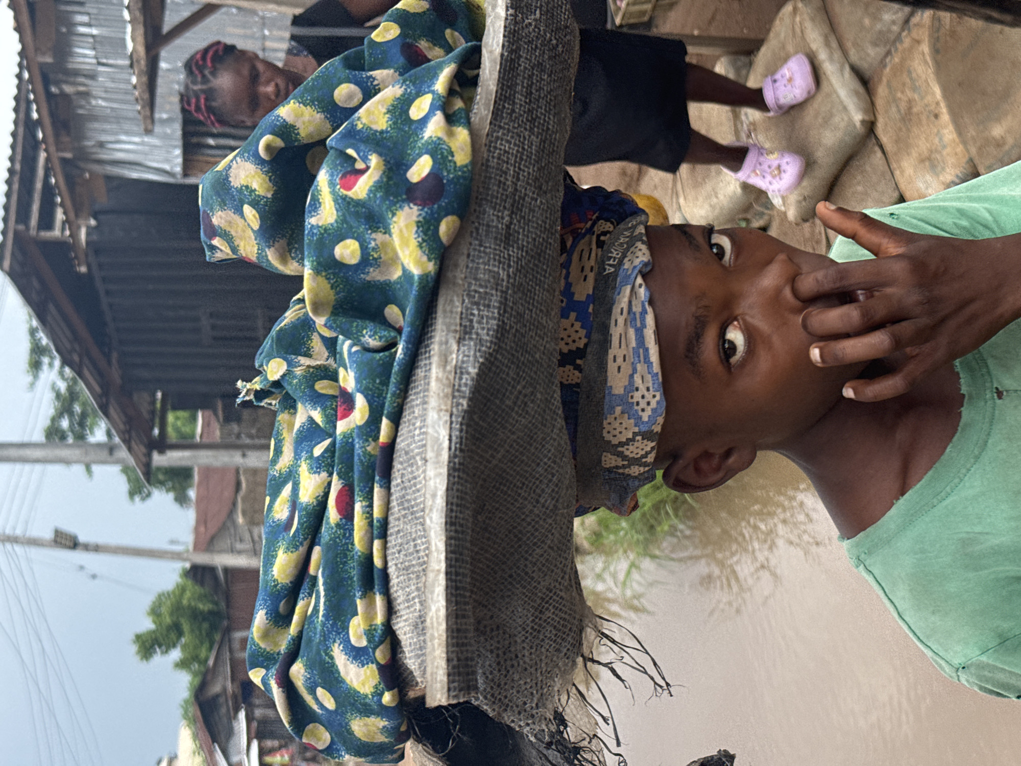 This outdoor portrait captures two individuals in what appears to be a community in Ihiagwa, Nigeria. In the foreground, a young person with dark skin wears a light green t-shirt and a patterned headscarf featuring blue, white, and brown geometric designs. This individual looks directly at the camera with wide eyes, and their left hand is raised towards their nose or mouth. They are carrying a substantial, wrapped load on their head, covered by a teal fabric adorned with yellow and red circular patterns, further encased in a coarse, grey mesh material.
In the mid-ground, partially obscured, another person is visible, characterized by reddish braided hair and a dark top. Their feet, clad in light purple Crocs, rest on large stones or concrete blocks.
The background reveals an outdoor setting featuring structures with corrugated metal roofs, indicative of a semi-urban or rural area. A significant portion of the background shows murky, brownish water, possibly suggesting a flooded area or a nearby body of water, with reflections of trees visible on its surface. Utility poles and power lines are discernible against an overcast sky, indicating daylight with diffused light. No explicit activity is depicted beyond the act of carrying the load and the direct gaze at the camera. Faint, indistinct text, possibly "MADARA," can be seen on the headscarf of the foreground subject.