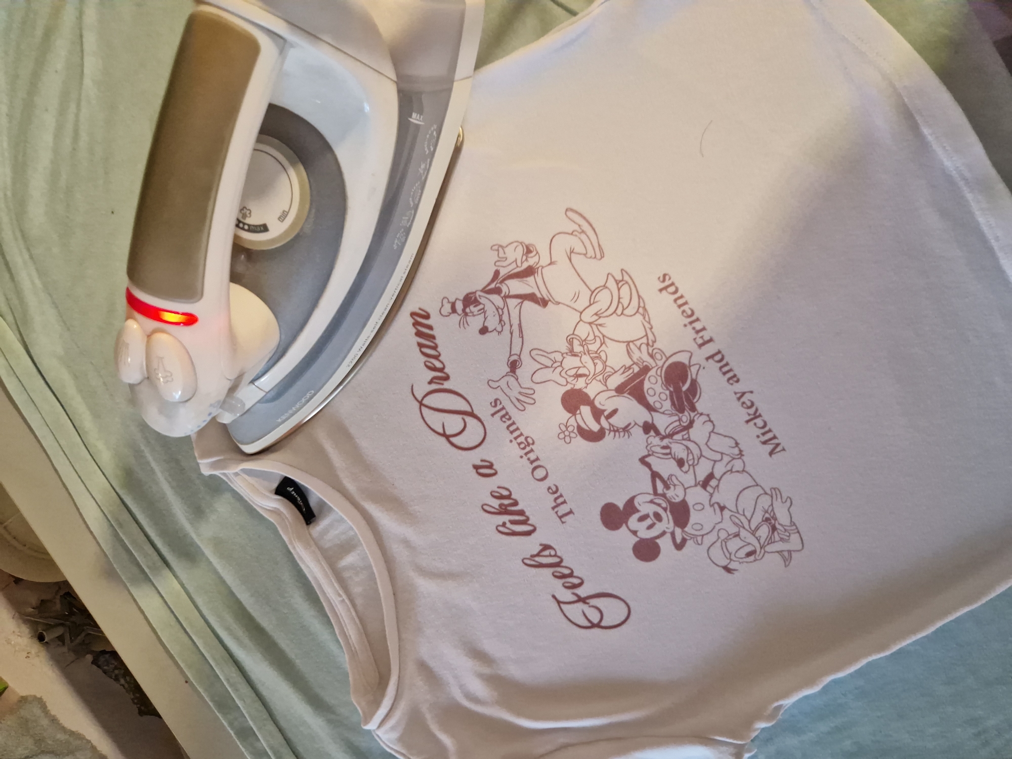 An off-white t-shirt with a dark pink graphic is laid out on a pale green surface, possibly a bed or ironing board. The graphic features several Disney characters, including Mickey Mouse, Minnie Mouse, Donald Duck, Daisy Duck, and Goofy, arranged in a cluster. Above the characters, the text "Feels like a Dream" is written in a cursive font, and below them, "The Originals" and "Mickey and Friends" are printed in a simpler font. A white and grey iron with a bronze grip is positioned on the t-shirt, with its red indicator light illuminated. The background is out of focus, showing some clutter. There is no information to determine the location in the city of Sofia, Bulgaria.
