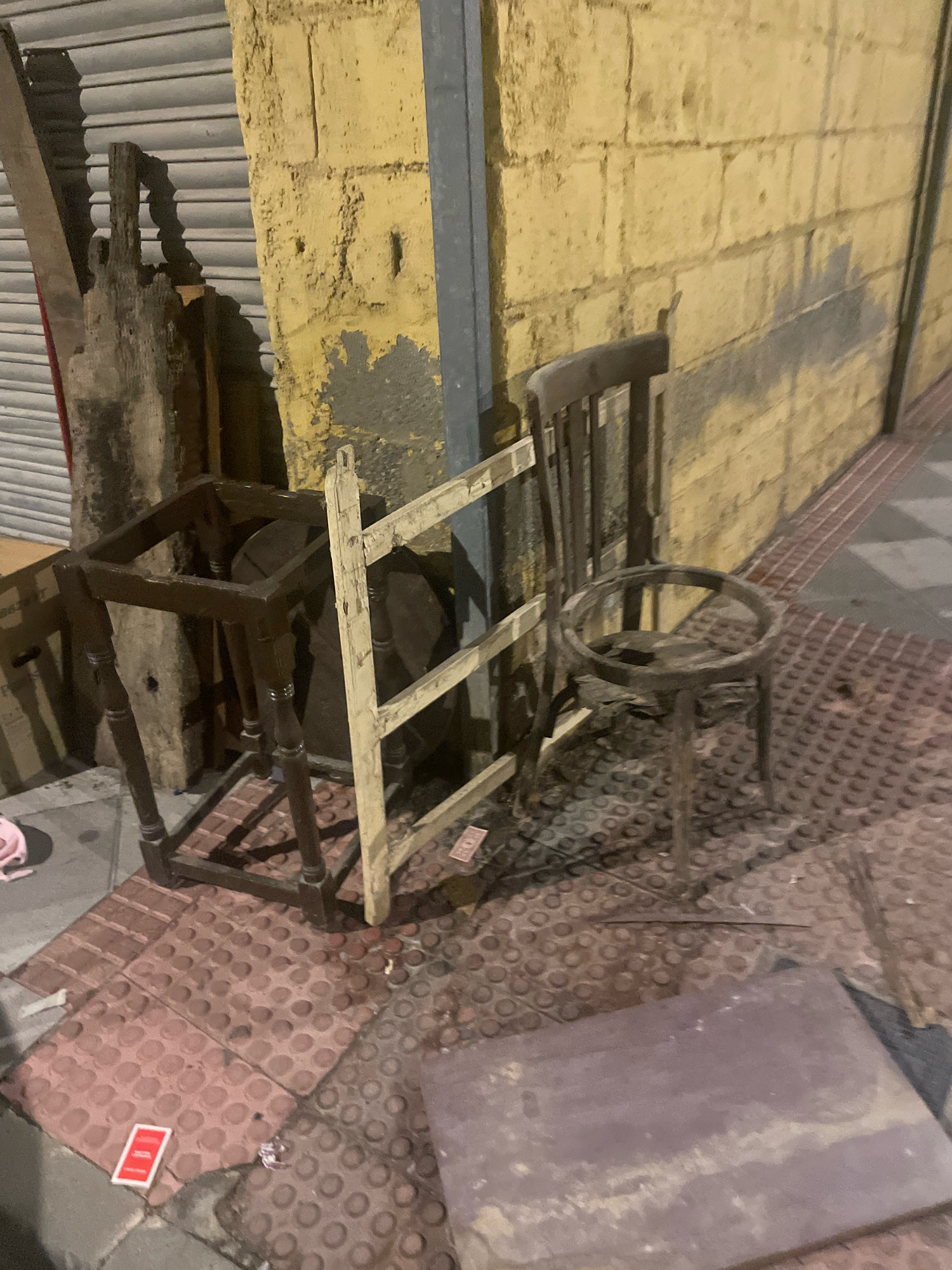 The image depicts an outdoor scene in Atarfe, Spain, with a focus on discarded furniture and debris on a sidewalk. In the foreground, a dark wooden table frame with four turned legs and a square top is overturned. Adjacent to it, a partially dismantled wooden structure with white painted slats and a frame lies on its side. To the right, a circular metal object, possibly a stool or a part of a larger item, stands on three thin legs. Behind these objects, a dark wooden chair with vertical slats on its back and seat is positioned against a textured yellow wall. The ground is paved with reddish-brown tactile paving tiles and appears wet and dirty, with scattered debris, including a red and white playing card and what looks like a piece of purple material. A metal roller shutter door is visible in the background to the left, behind some wooden planks. The lighting suggests it is nighttime or a dimly lit indoor space.