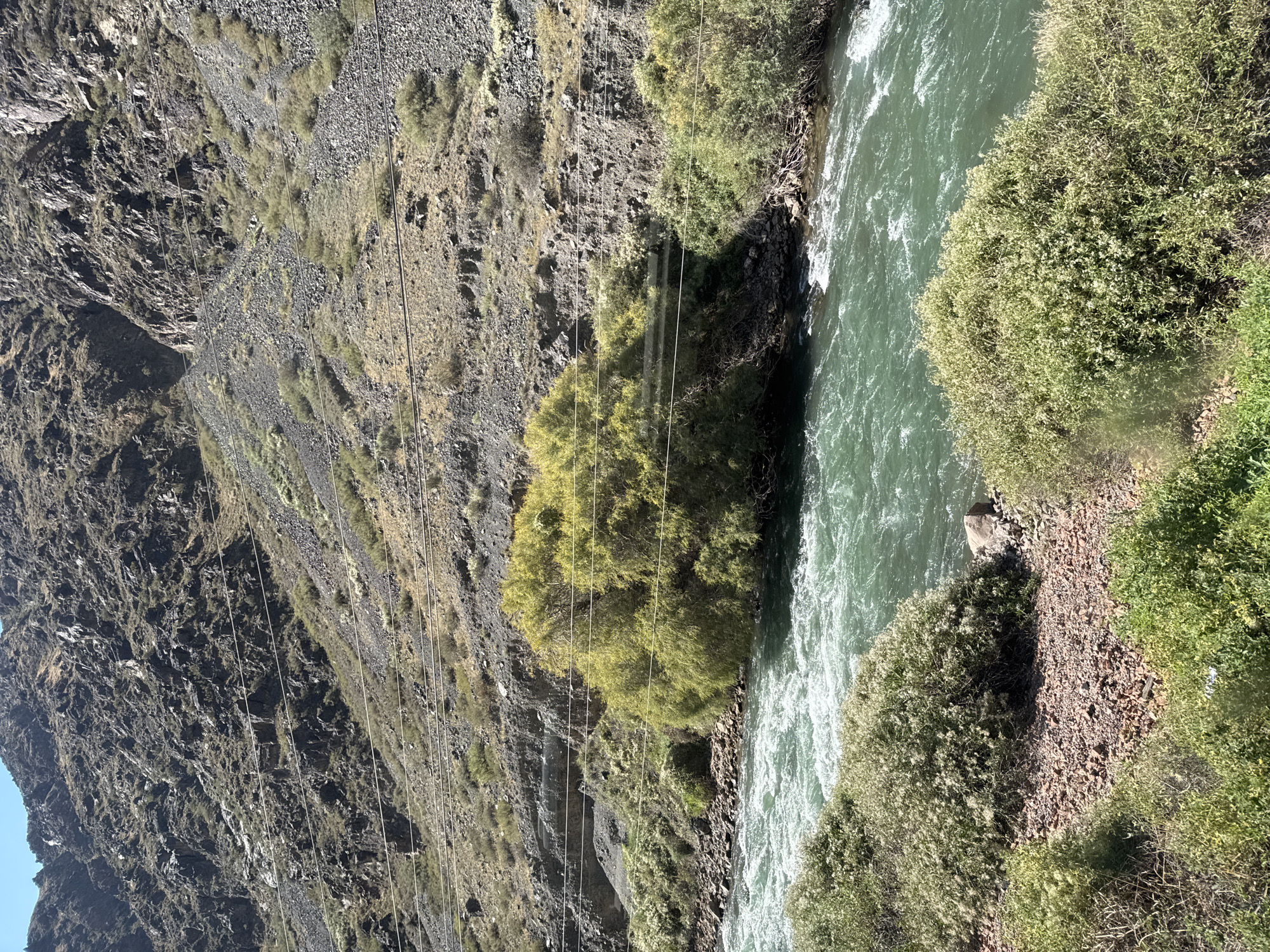 The image depicts a rugged mountain landscape with a flowing river. The terrain is steep and rocky, covered with sparse vegetation and gravel. Several parallel power lines stretch across the mountainside, suggesting human infrastructure. A dense cluster of green trees with lighter foliage stands prominently near the riverbank. The river itself is a vibrant turquoise, with visible currents and ripples. The foreground is characterized by rocky ground and bushy green plants. The sky above is clear and blue. The scene appears to be in a natural, undeveloped area, possibly a mountain valley or gorge. The location is identified as a specific region within Kyrgyzstan, though the exact city or town is not specified.
