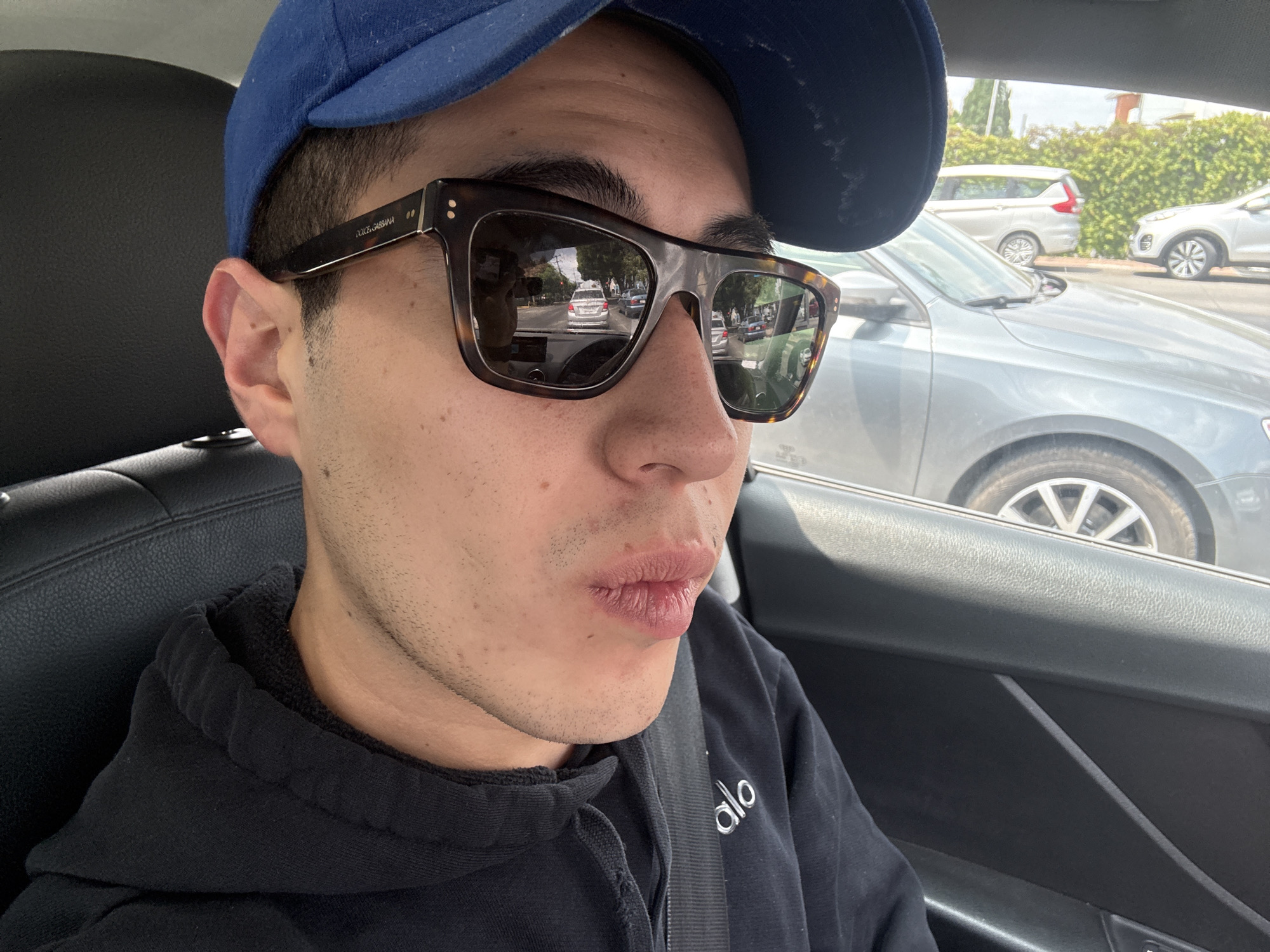 A person is in a car, wearing a blue baseball cap, Dolce & Gabbana sunglasses, and a black hoodie with an "Alo" logo. The person appears to be driving or in the front passenger seat. Outside the car, vehicles and foliage are visible, suggesting an urban or suburban environment. The location is not Puebla, Mexico.