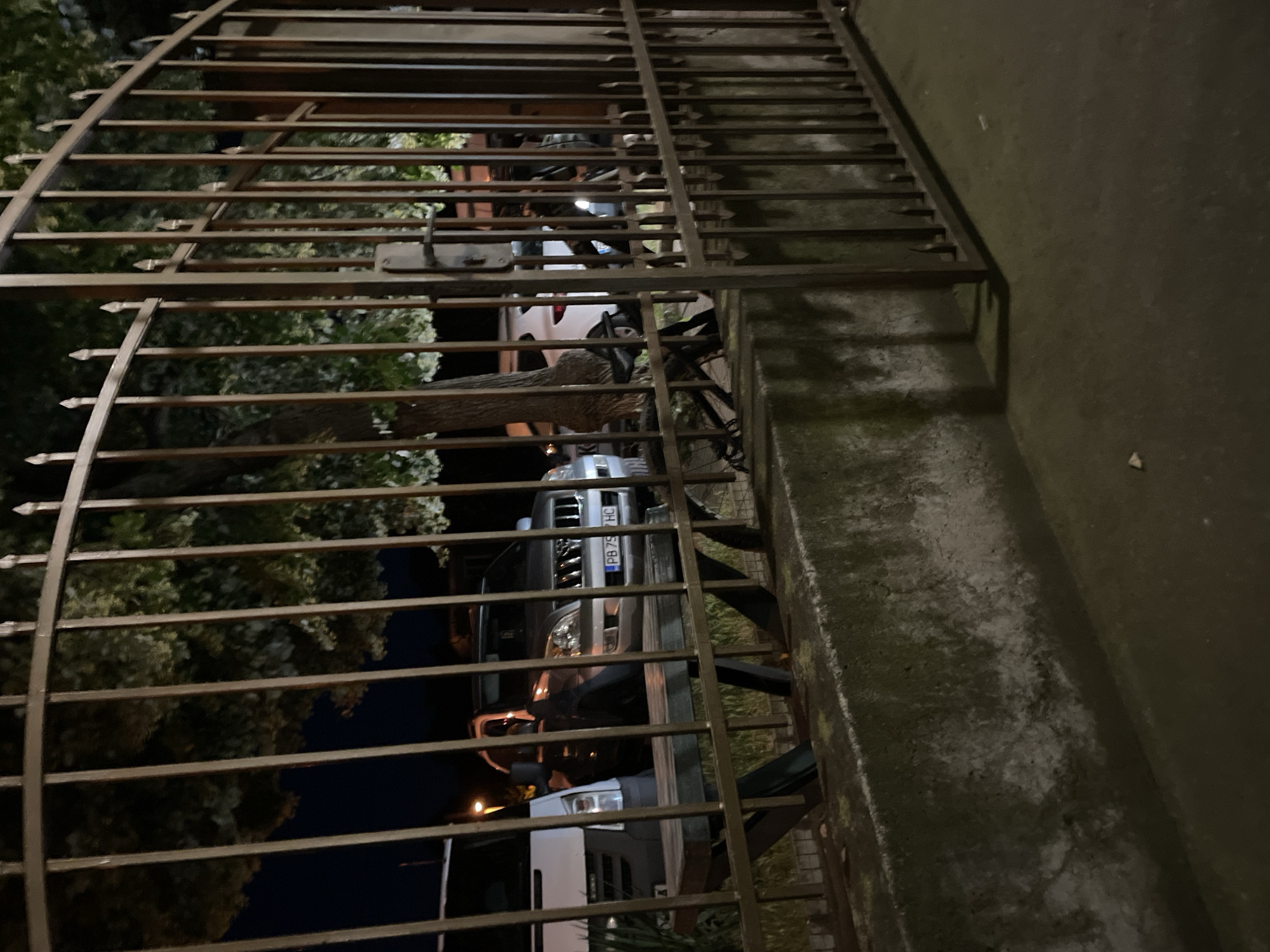 A nighttime outdoor scene in Muldava, Bulgaria, features a dark, wrought-iron gate with pointed finials. Through the gate, a concrete retaining wall is visible, covered in patches of moss. Behind the wall, a wooden picnic table with metal legs is partially obscured by the gate. A silver SUV with the license plate "PB 75 HC" is parked behind the table. To the left of the SUV, a white van is also parked. Trees with dense foliage are visible in the background, with some leaves illuminated by light sources not visible in the frame. The ground in the foreground is a dark, asphalt-like surface.