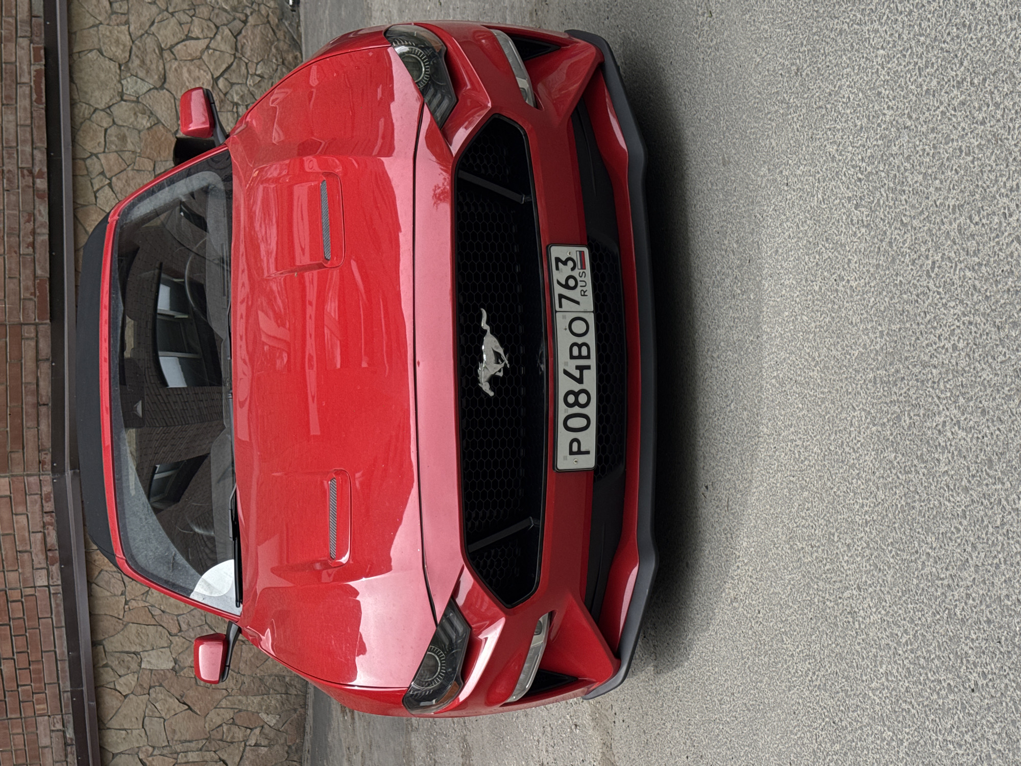 The image shows a red Ford Mustang parked on a gray pavement in an urban environment. The car is angled slightly, presenting a view of the front and side. The car has a Russian license plate "P084BO 763 RUS". To the left, a brick and stone wall is visible. The location is Unknown, Unknown.