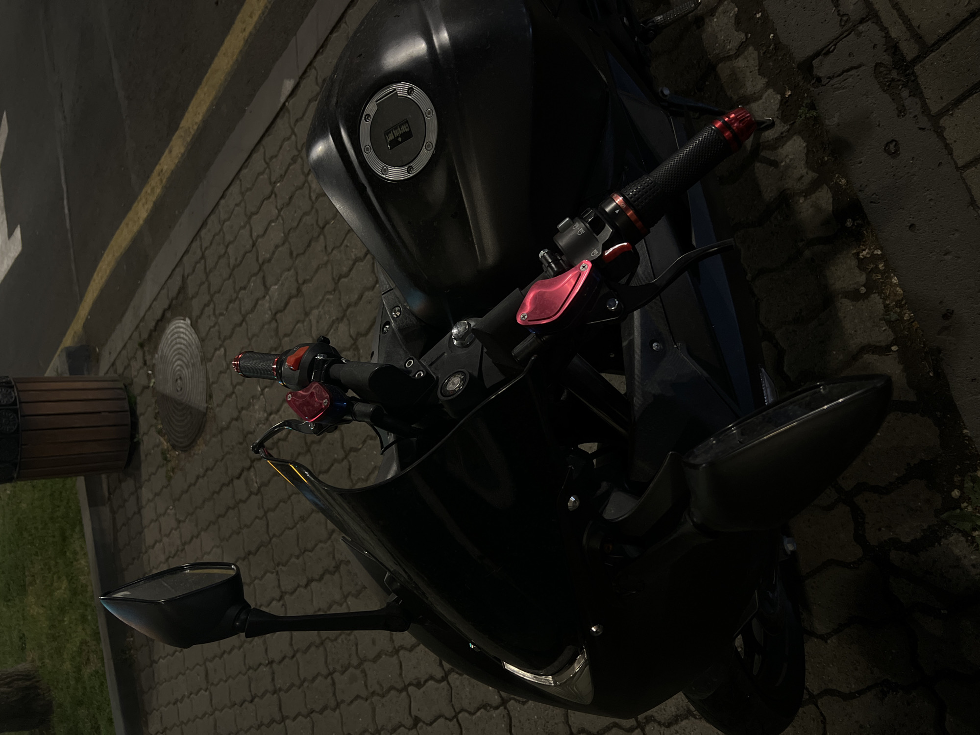 The image captures a close-up, overhead view of a black motorcycle parked on a cobblestone surface. The motorcycle's handlebars are equipped with black and red grips, and a red reservoir is visible. The fuel tank is prominently featured, with a gas cap bearing legible text. The motorcycle is parked near a curb marked with a yellow line, and a wooden cylindrical object is partially visible in the background. The setting appears to be an urban environment at night, with street lighting casting shadows and highlighting the textures of the cobblestone and the motorcycle's surfaces.