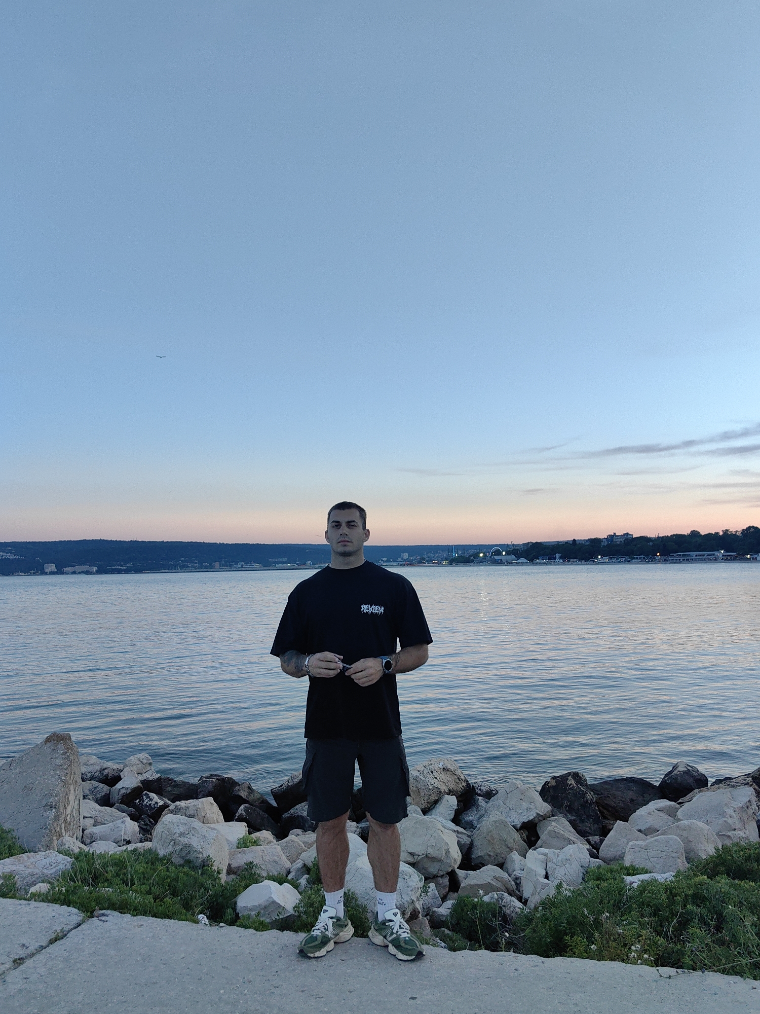 A full-body, eye-level outdoor shot captures a young man standing on a concrete path next to a rocky shoreline. The man is facing the camera, holding sunglasses in his hands. He is wearing a black t-shirt, dark shorts, white socks, and green and white athletic shoes. Behind him, a calm body of water stretches to the horizon, where a city skyline is visible under a twilight sky. The sky transitions from a pale blue at the top to soft hues of pink and orange near the horizon. The water has gentle ripples, reflecting the colors of the sky. In the upper left quadrant of the sky, a small airplane is visible. The rocky shoreline is covered with patches of green vegetation. The overall scene suggests an evening setting by the water in a coastal urban area.