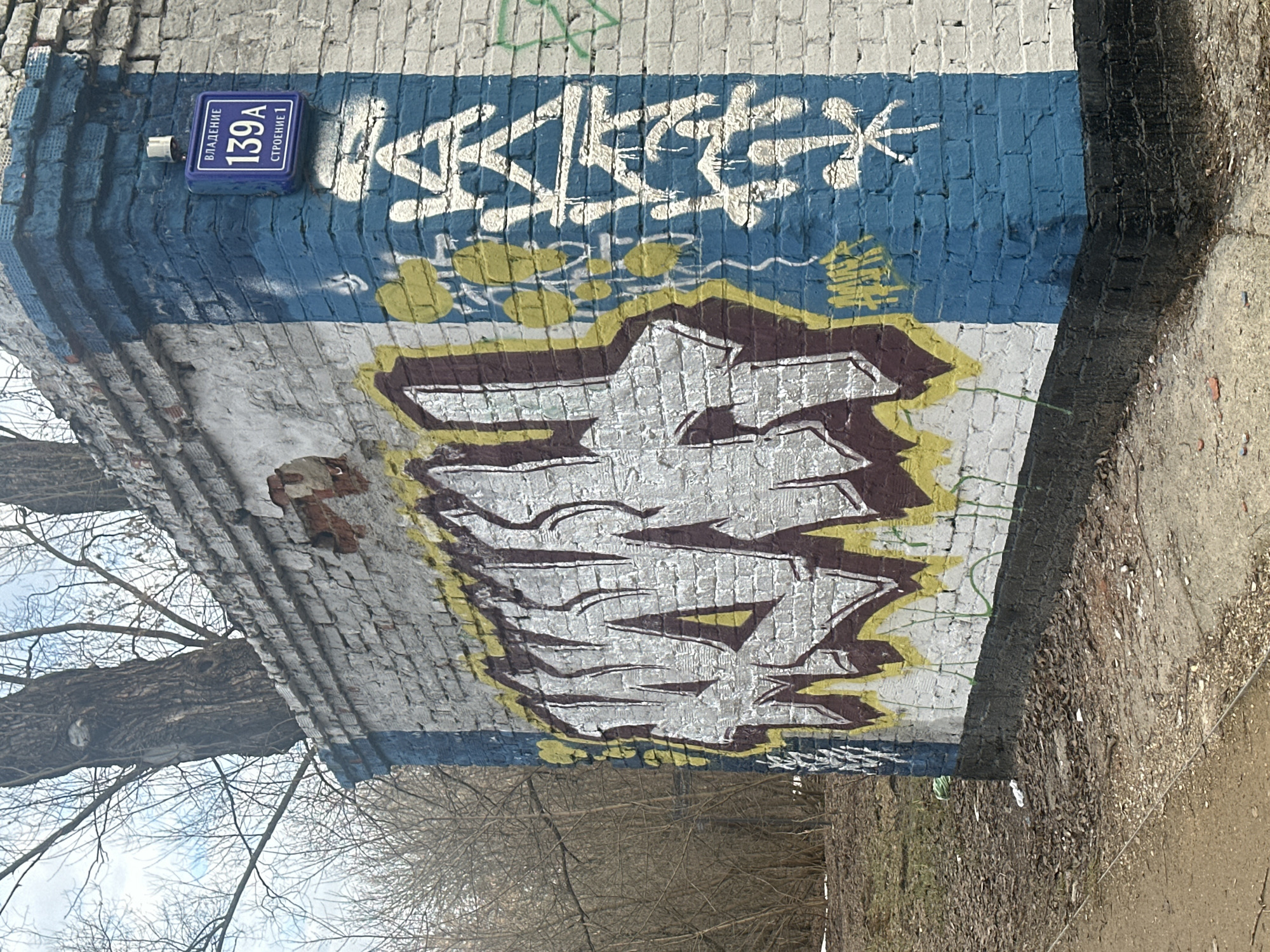 A brick wall with graffiti and a street sign is located outdoors. The street sign reads "VLADENIE 139A STROENIE 1," which translates to "Property 139A Building 1," indicating a location in Moscow, Russia. The graffiti includes a white tag on a blue background at the top, with yellow accents, and a large, outlined tag in white and brown. The wall shows signs of age and disrepair. A bare tree is visible to the left. To the right is the edge of a sidewalk. The ground near the wall is covered with dried leaves and debris.
