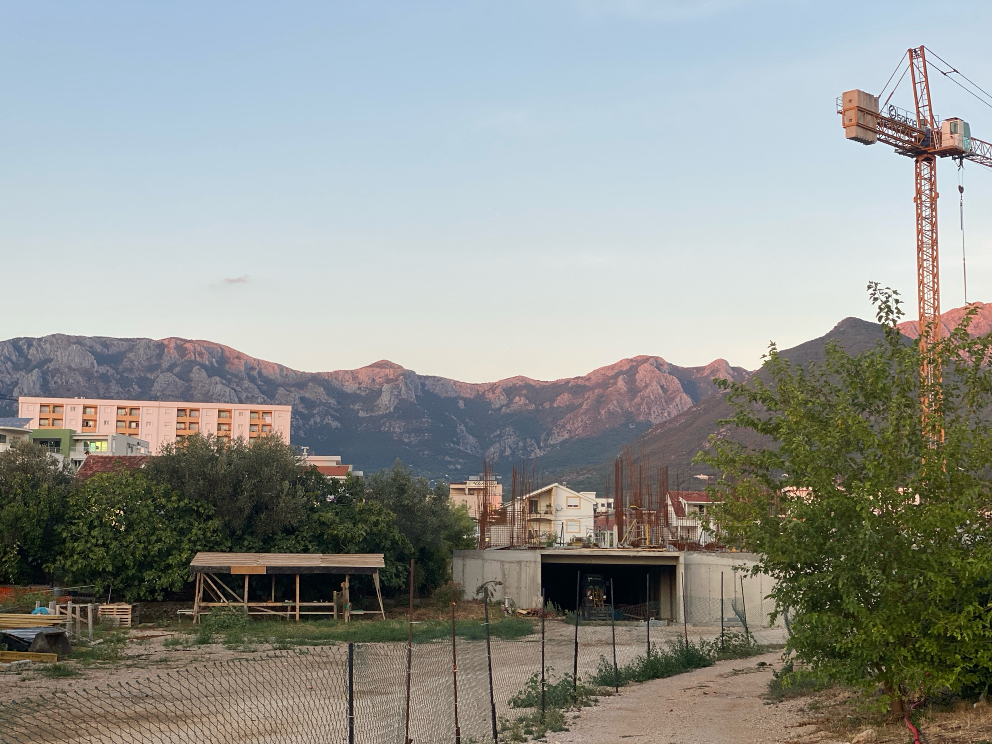 This is a landscape photograph taken in the city of Bar, Montenegro, during what appears to be the late afternoon or early evening. The sky is clear and a pale blue, transitioning to a lighter hue near the horizon. Dominating the background are rugged, mountainous peaks, their slopes catching the warm, golden light of the setting sun.
In the middle ground, a long, multi-story apartment building with a light-colored facade and numerous balconies is visible. To its left, a portion of another building, possibly under construction or renovation, can be seen with green accents. A large construction crane, painted orange, stands prominently in the upper right quadrant of the image, its jib extended over the scene.
The foreground features a construction site. A concrete structure, likely the foundation or lower level of a building, is partially visible with rebar protruding. A temporary wooden shelter with a sloped roof stands to the left of this structure. Various construction materials, including wooden planks and pallets, are scattered around the site. A chain-link fence encloses a portion of the area, with a dirt path running alongside it. Lush green trees and foliage are present throughout the scene, with a particularly dense tree in the right foreground partially obscuring the view of the construction crane. The overall impression is one of ongoing development against a backdrop of natural mountain beauty.