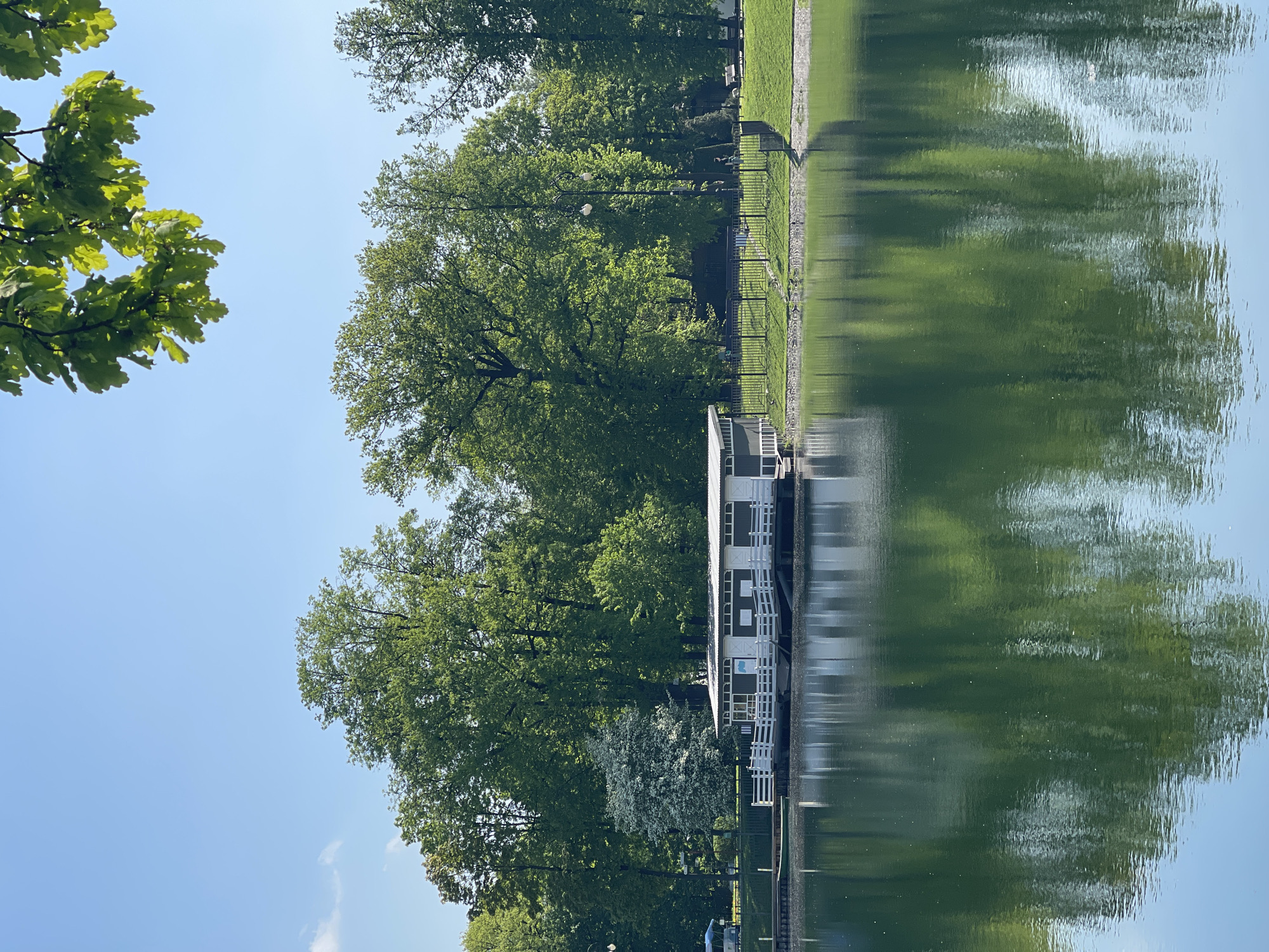 The image depicts a body of water reflecting a scene on its shore. The shore features lush greenery, including trees and grass. A building with a white facade is situated near the water's edge, its reflection visible in the water. The scene is set against a clear blue sky. The location appears to be a park or recreational area, but its specific city and region, Unknown, Unknown, are indeterminate.