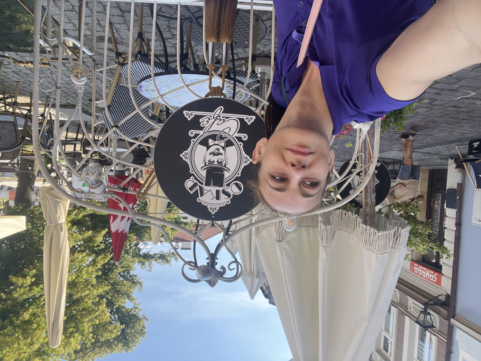 This is a selfie taken outdoors at a cafe. A woman is prominently featured in the foreground, wearing a purple shirt and a pink strap across her shoulder. She is looking directly at the camera with a neutral expression. Behind her, the cafe setting is visible. There is a black circular sign with a white illustration of a man in a hat and the word "Cafe" written in stylized script. The sign hangs from a decorative white metal structure. Various cafe tables and chairs with black and white striped cushions are scattered around. Several large, cream-colored umbrellas with fringed edges are deployed, suggesting it's a sunny day, possibly late morning or afternoon. One umbrella is covered with a beige fabric. In the background, trees with green foliage are visible, as well as buildings with European-style architecture. A red umbrella with white designs is also seen. The ground appears to be paved with cobblestones. The overall atmosphere seems relaxed and casual, typical of an outdoor cafe setting. Visible text includes "Cafe" on the sign and some blurred text on a red sign in the background.