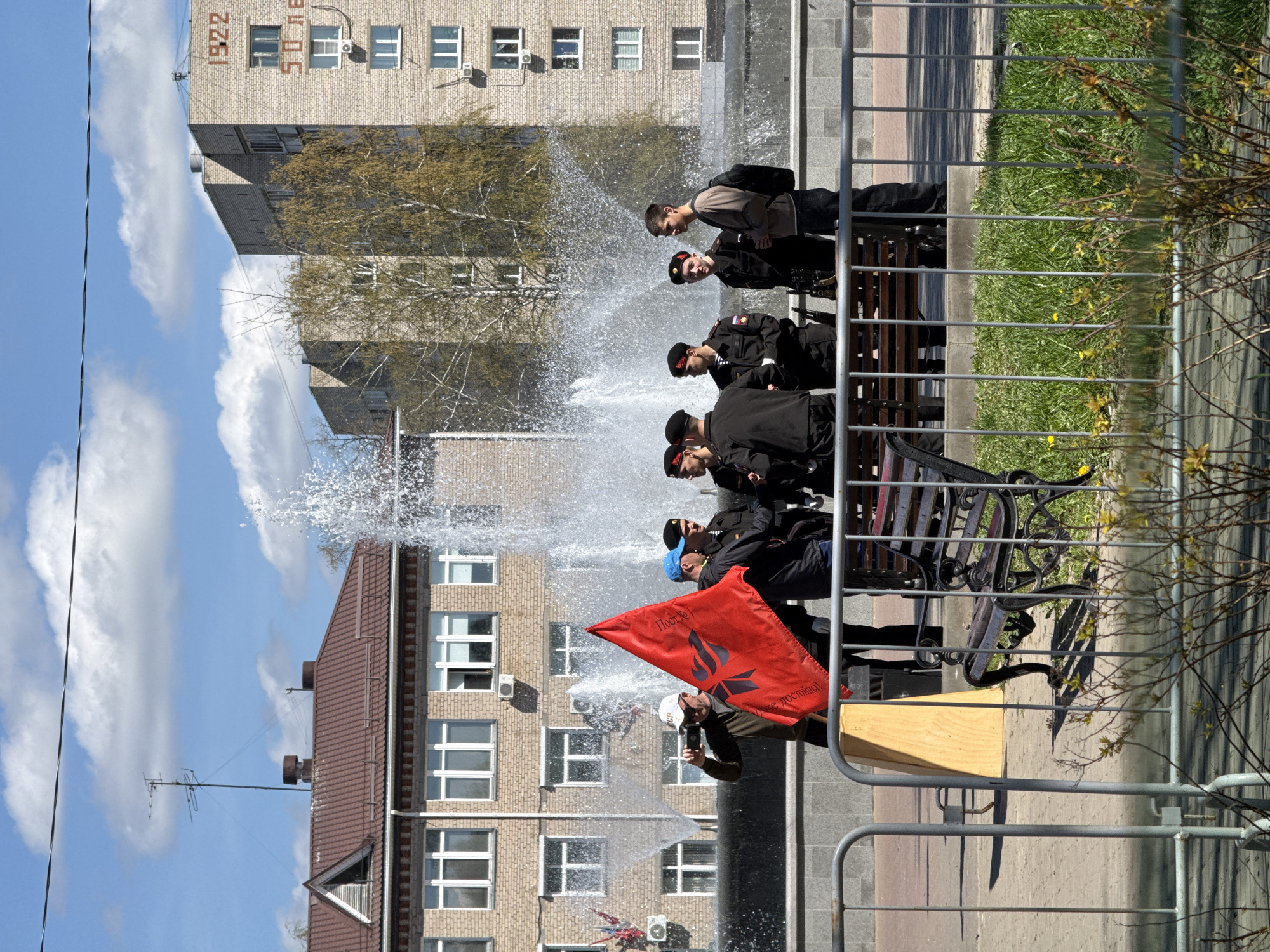 A scene is captured near a fountain in what appears to be a city setting. Several individuals dressed in black uniforms with peaked caps sit on a bench next to a metal railing. A person in civilian clothing stands slightly in front of the group, holding a bright red flag with the inscription "Postoy za sebya - bud dostoynym!" (Stand up for yourself - be worthy!).
Behind the figures, a fountain sprays water. The backdrop includes a multi-story building with light-colored brickwork, and a rooftop with a brown gable, possibly part of another building, is visible below. In the background, there is a building with the numbers "1922" and the words "50 let" (50 years) on its facade.
Based on this information and the presence of the fountain with the "50 years" sign, it is likely that this photograph was taken at the fountain on Lenin Street in Chaikovsky, Russia, near the building with the inscriptions.