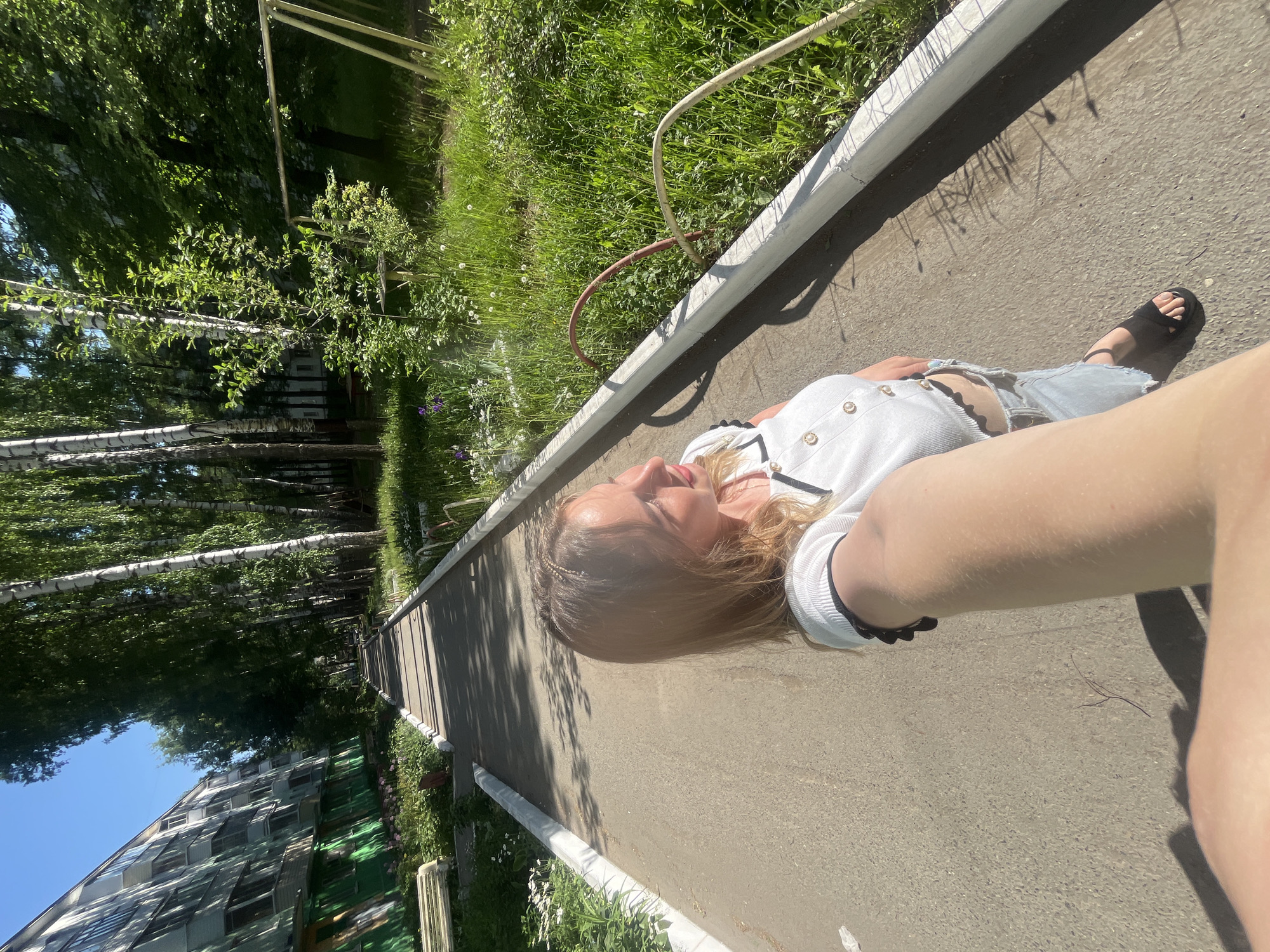 A woman with blond hair is lying on a paved path next to a grassy area, possibly in the city of Izhevsk, Russia. The woman is wearing a light-colored shirt, light-colored denim shorts, and black sandals. She is positioned at an angle, facing towards the left side of the frame. Her arm is extended towards the right, presumably holding a camera to take a selfie. The path is bordered by a concrete barrier. In the background, there are trees, including birch trees, and a building with green accents. A hose is visible near the edge of the path. The scene is brightly lit, suggesting it is daytime.